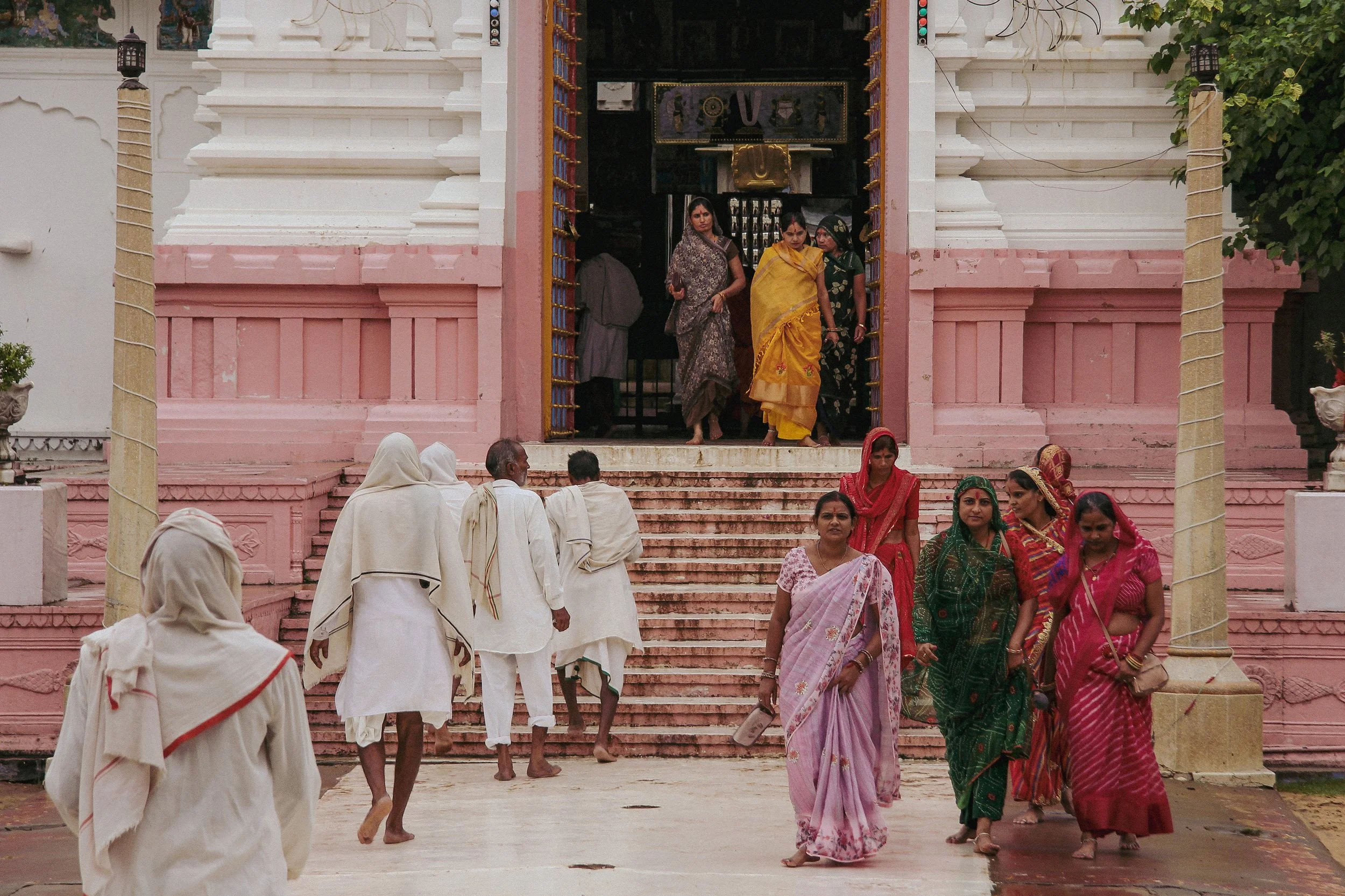 Group of women wearing colorful sarees and men in white clothing walking up steps towards a Hindu temple entrance, with some women entering the temple.