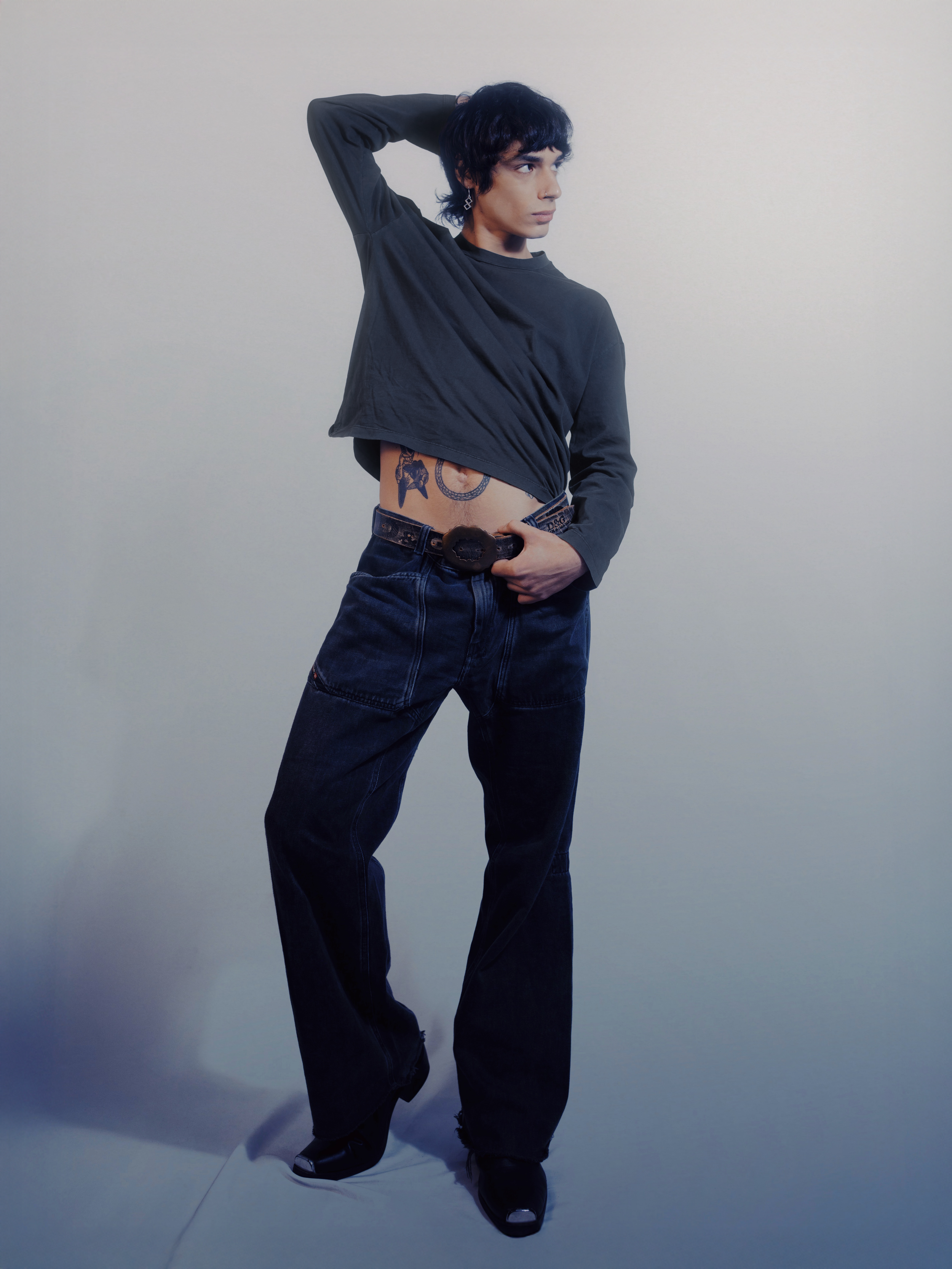 A person with short black hair, wearing a loose black long-sleeve shirt, baggy black jeans, and black shoes, standing against a plain light gray background with one hand behind their head and the other resting on their hip, revealing tattoos on their