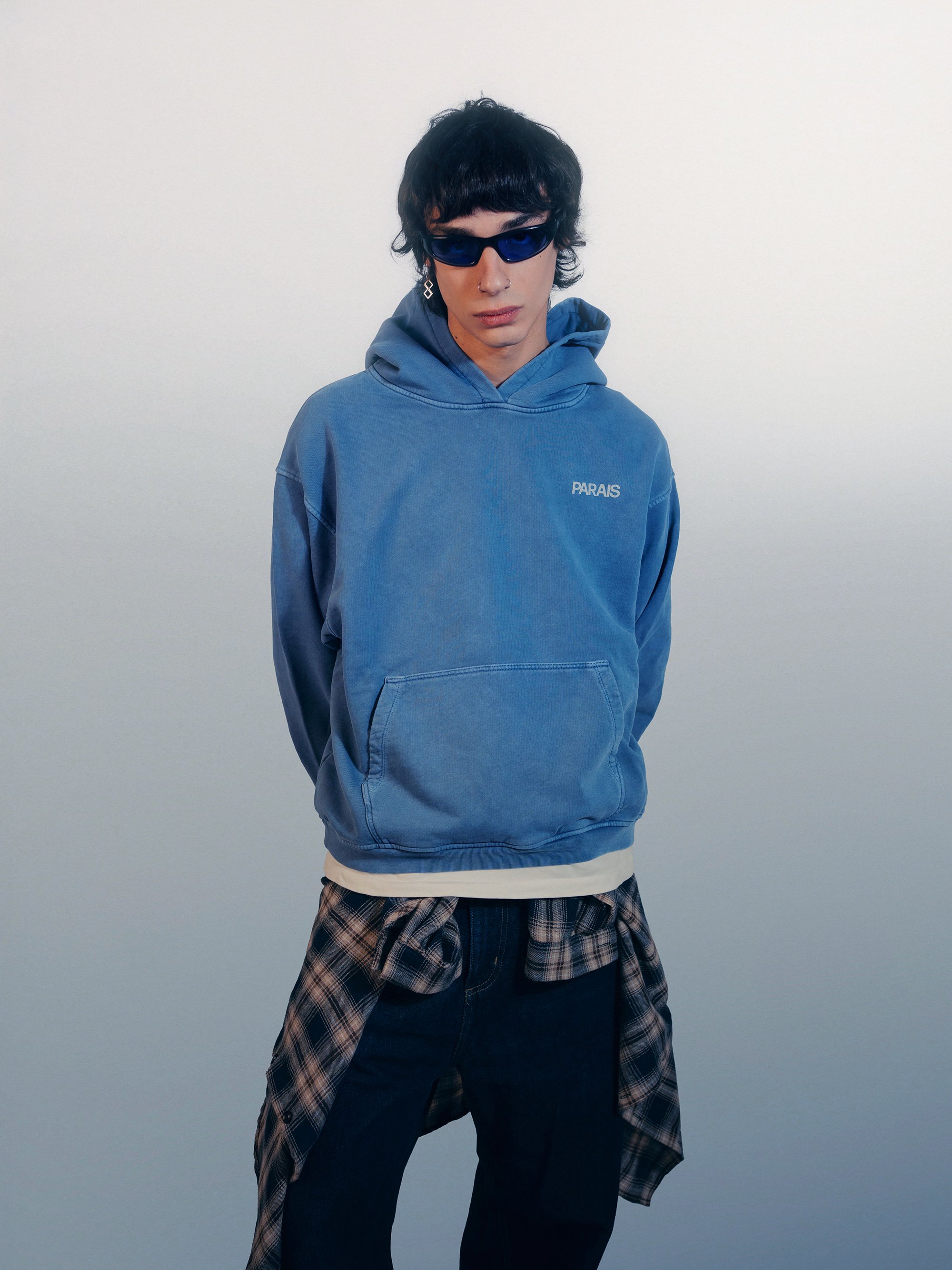 A young man with curly black hair wearing dark sunglasses, a blue hoodie with 'PARAIS' embroidered on it, dark pants, and a plaid shirt tied around his waist, standing against a plain white background.