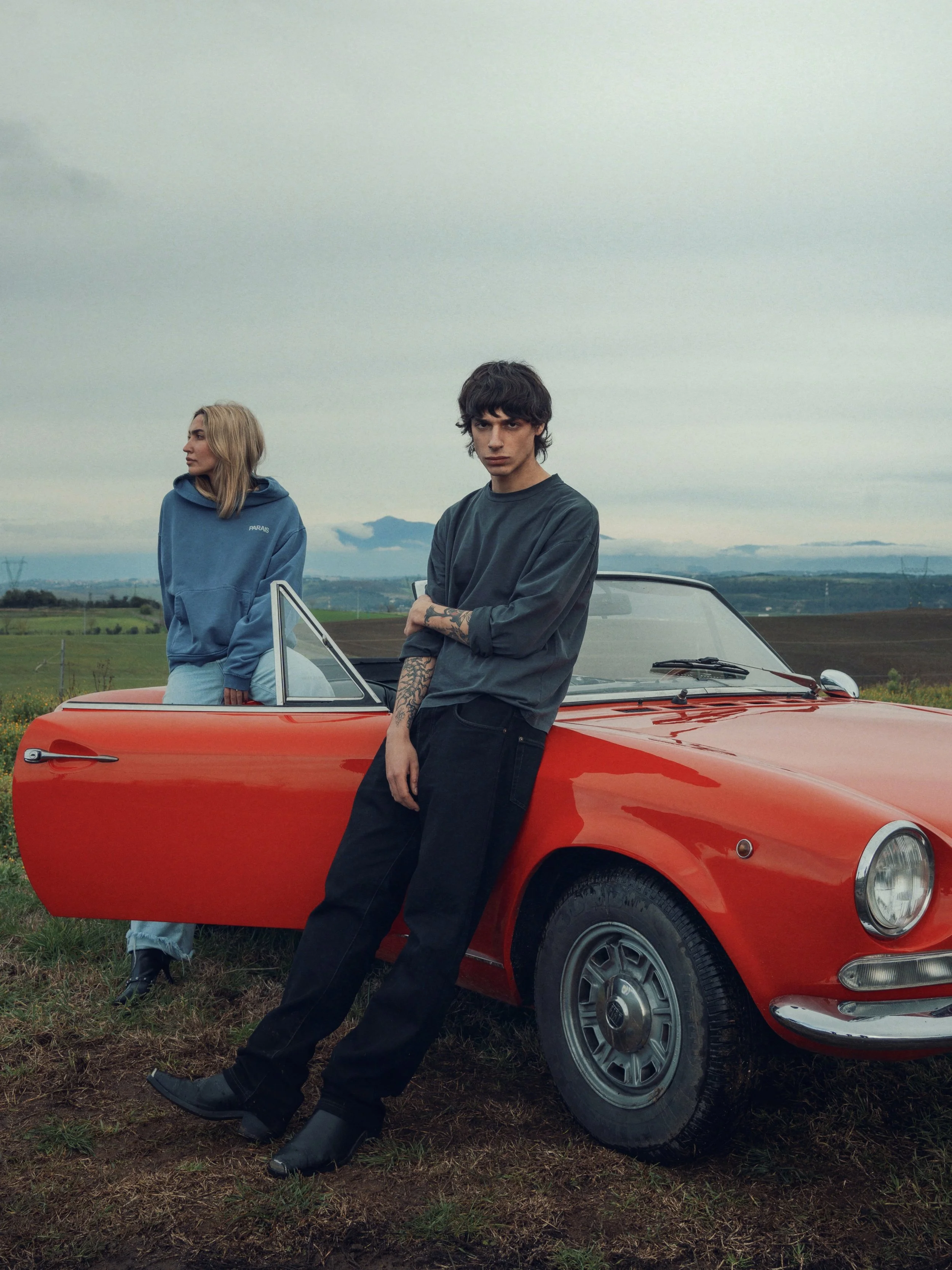 A young man leaning against a red vintage sports car with a woman standing inside the car's open door in a rural field under a cloudy sky.