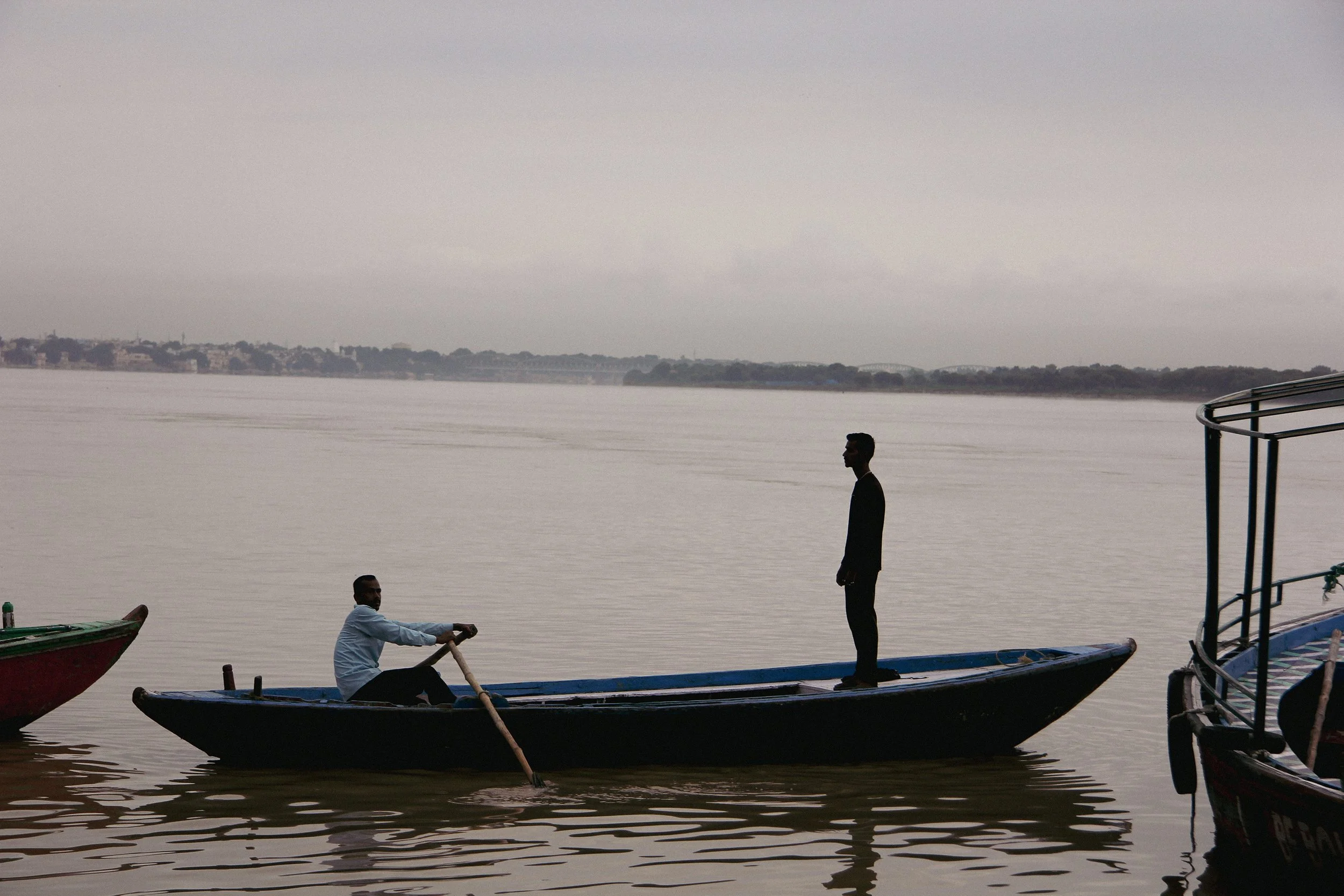 Two men on a boat on a calm body of water with a shoreline and buildings in the background, one standing and one sitting.