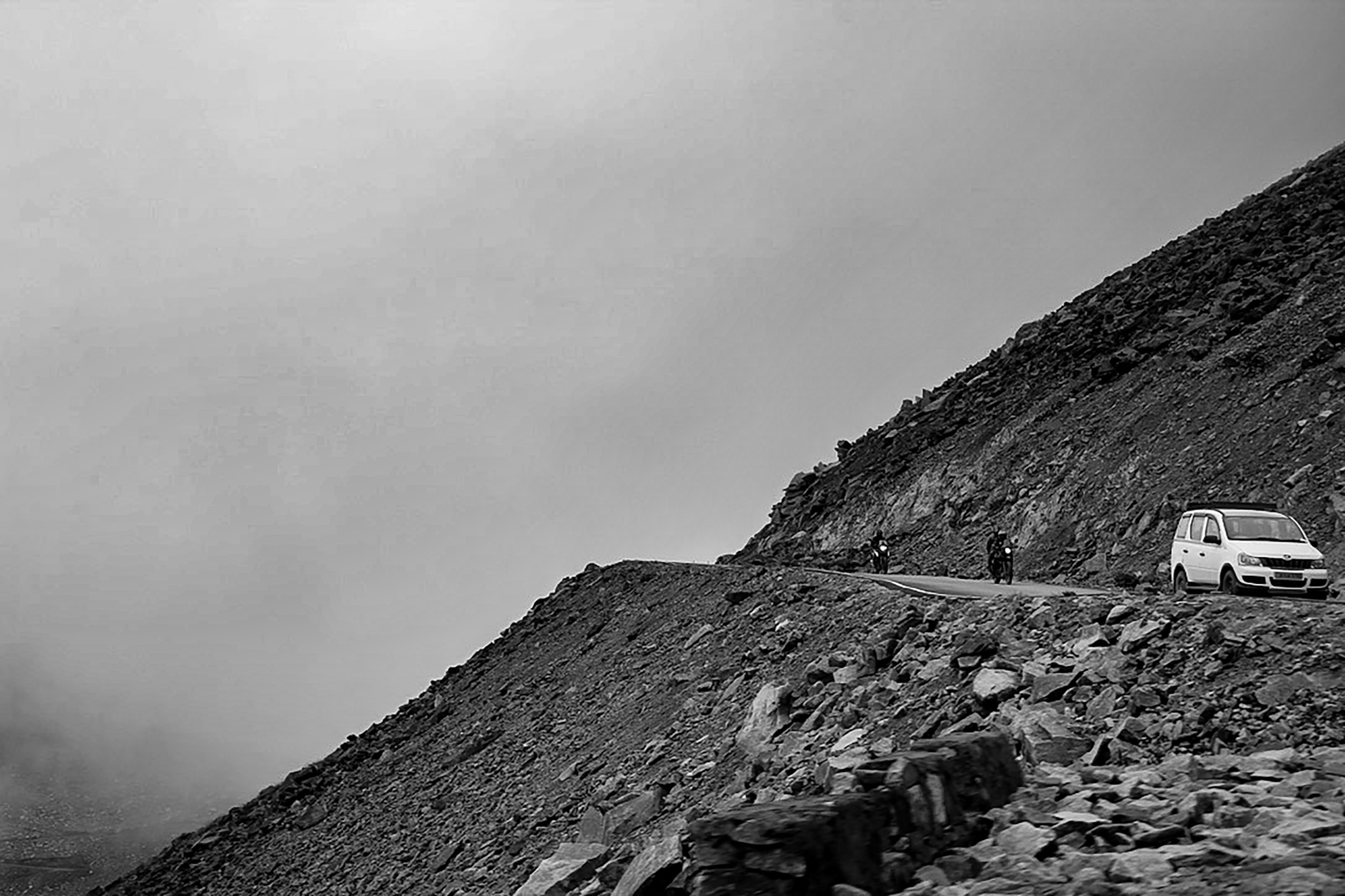 A black and white photo of a narrow mountain road with a white van and two motorcyclists riding along the rocky, steep hillside.