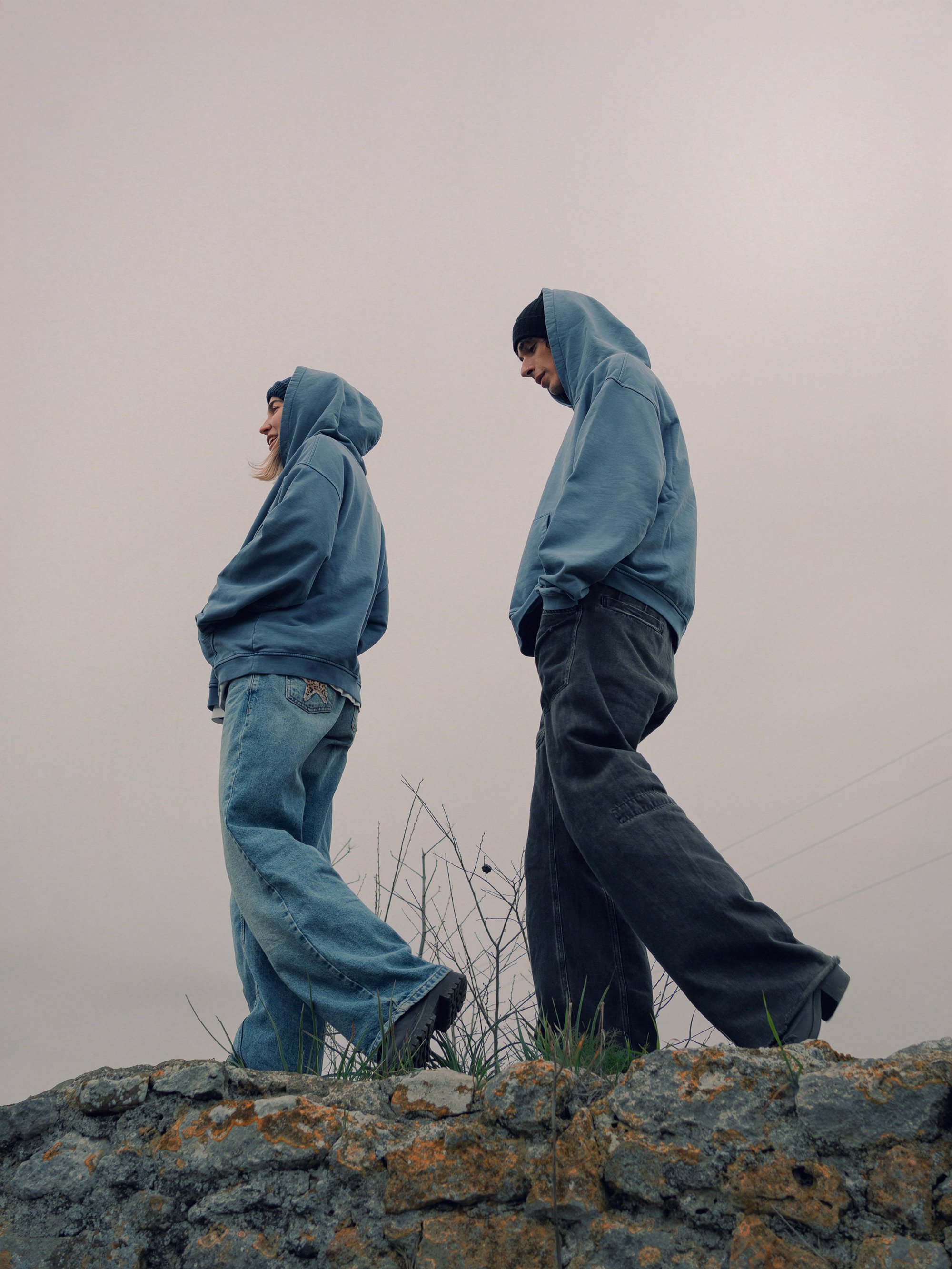 Two young people in hoodies and jeans standing on a rocky surface outdoors, facing downward with their heads bowed, against a cloudy sky.