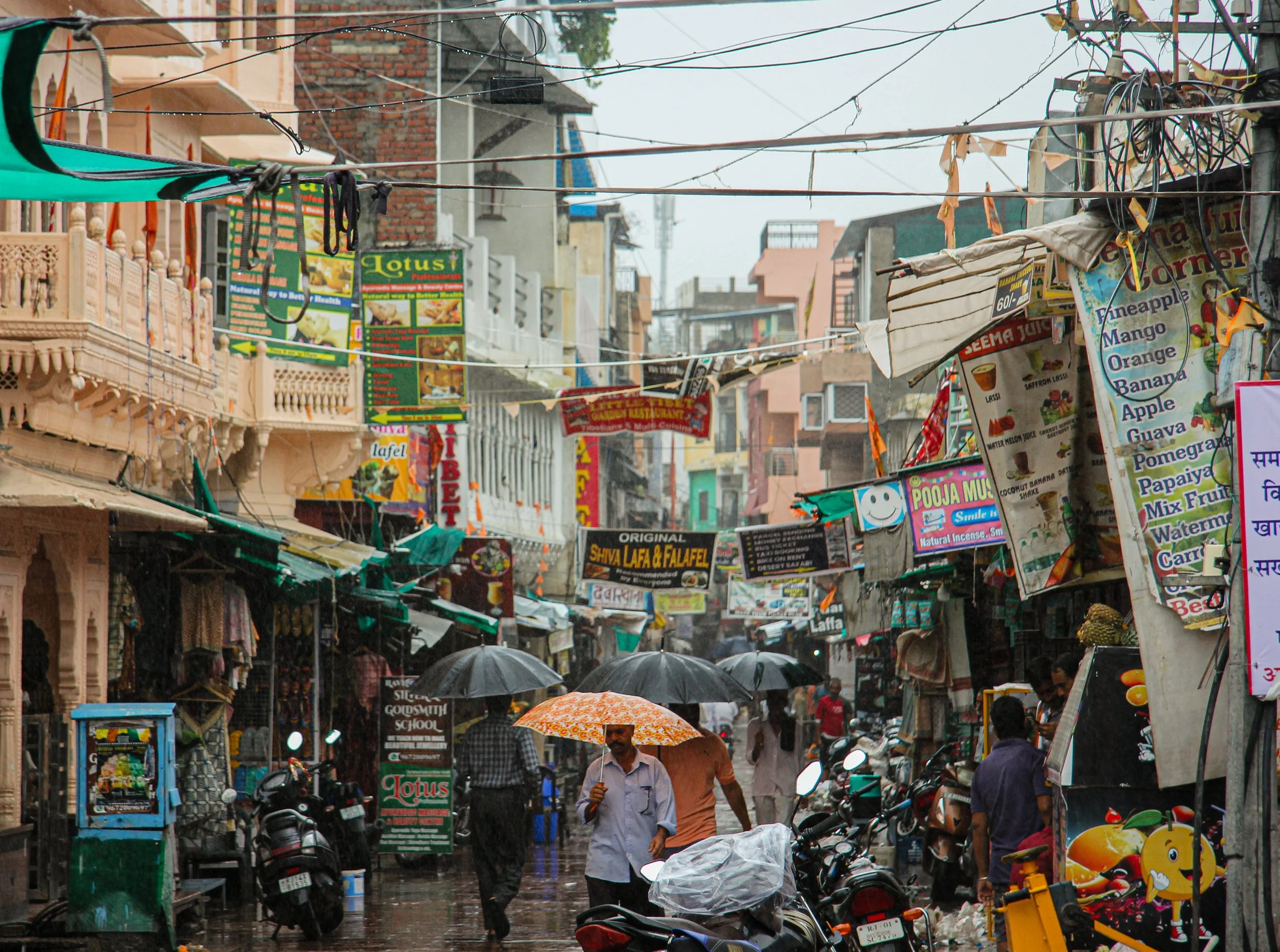 A busy street scene in an Asian city on a rainy day, with people walking under umbrellas, motorcycles parked along the sides, and colorful signs and shop signs hanging across the street.