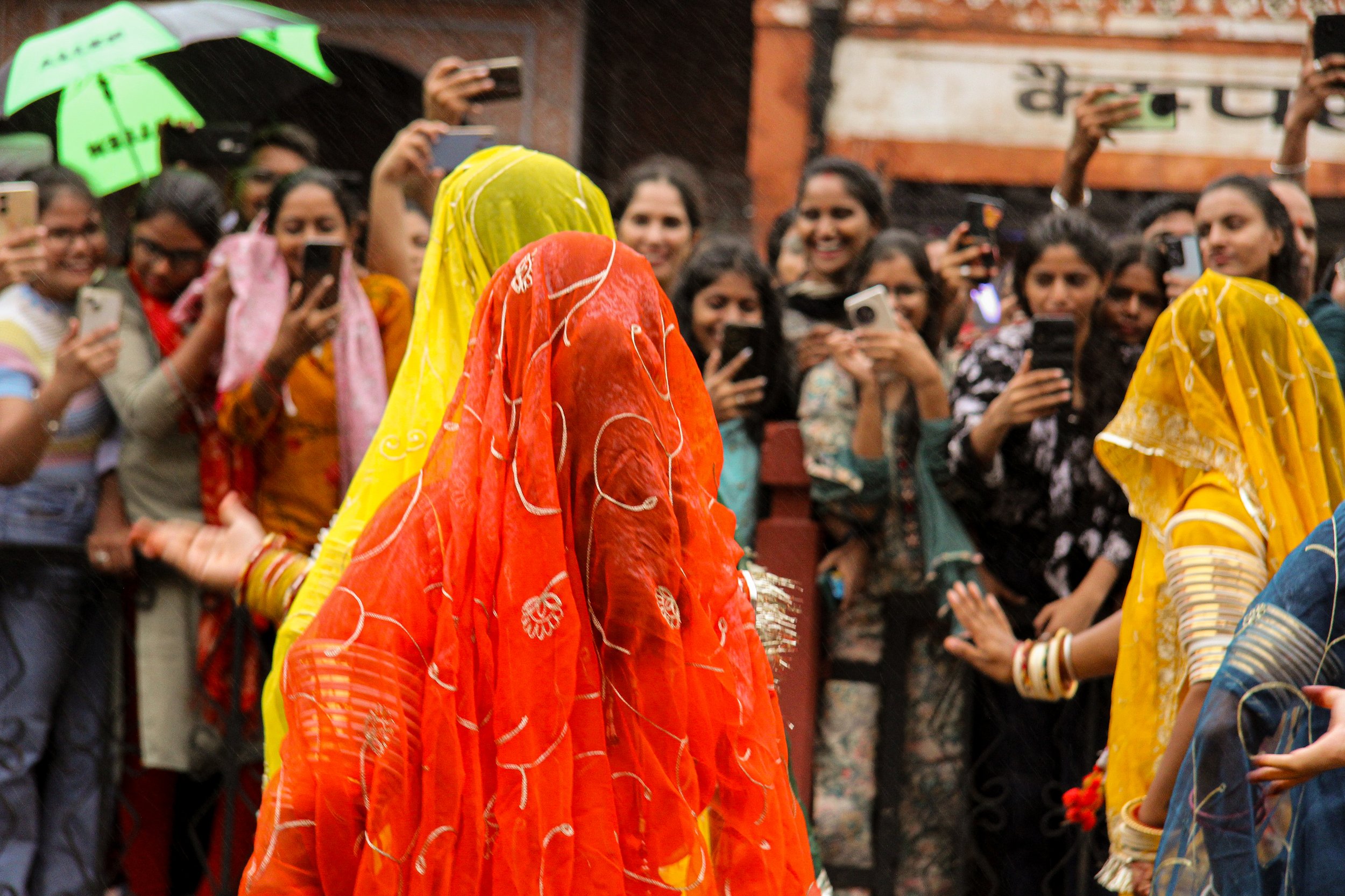 A group of women in colorful traditional Indian sarees, with some wearing bangles, taking photos and videos of women in bright orange, yellow, and blue sarees with rain in the background.