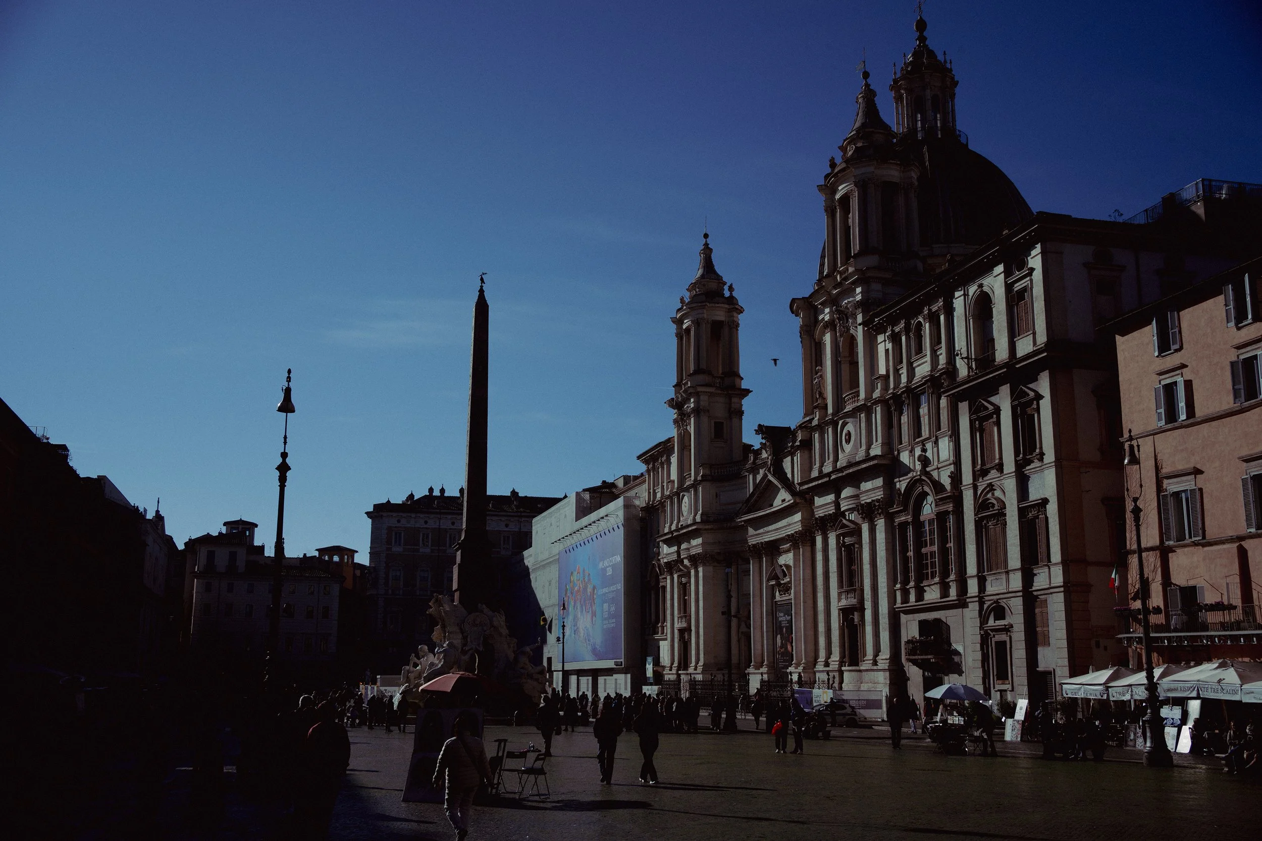 Sunset view of a historic baroque-style church with domes and towers, crowded piazza with people, street vendors, and an obelisk in the center, in Rome, Italy.