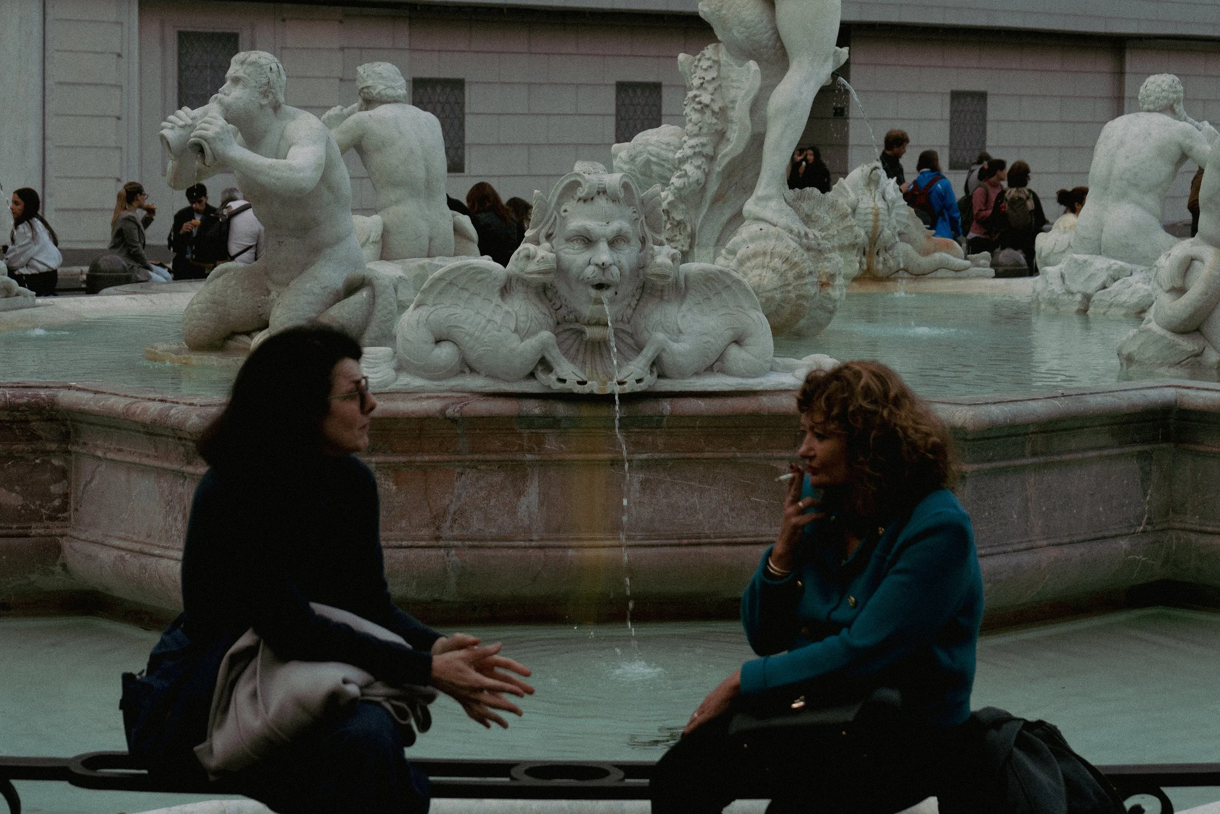Two women sitting and talking in front of a marble fountain with sculptures of mythological creatures, in a public plaza surrounded by people.
