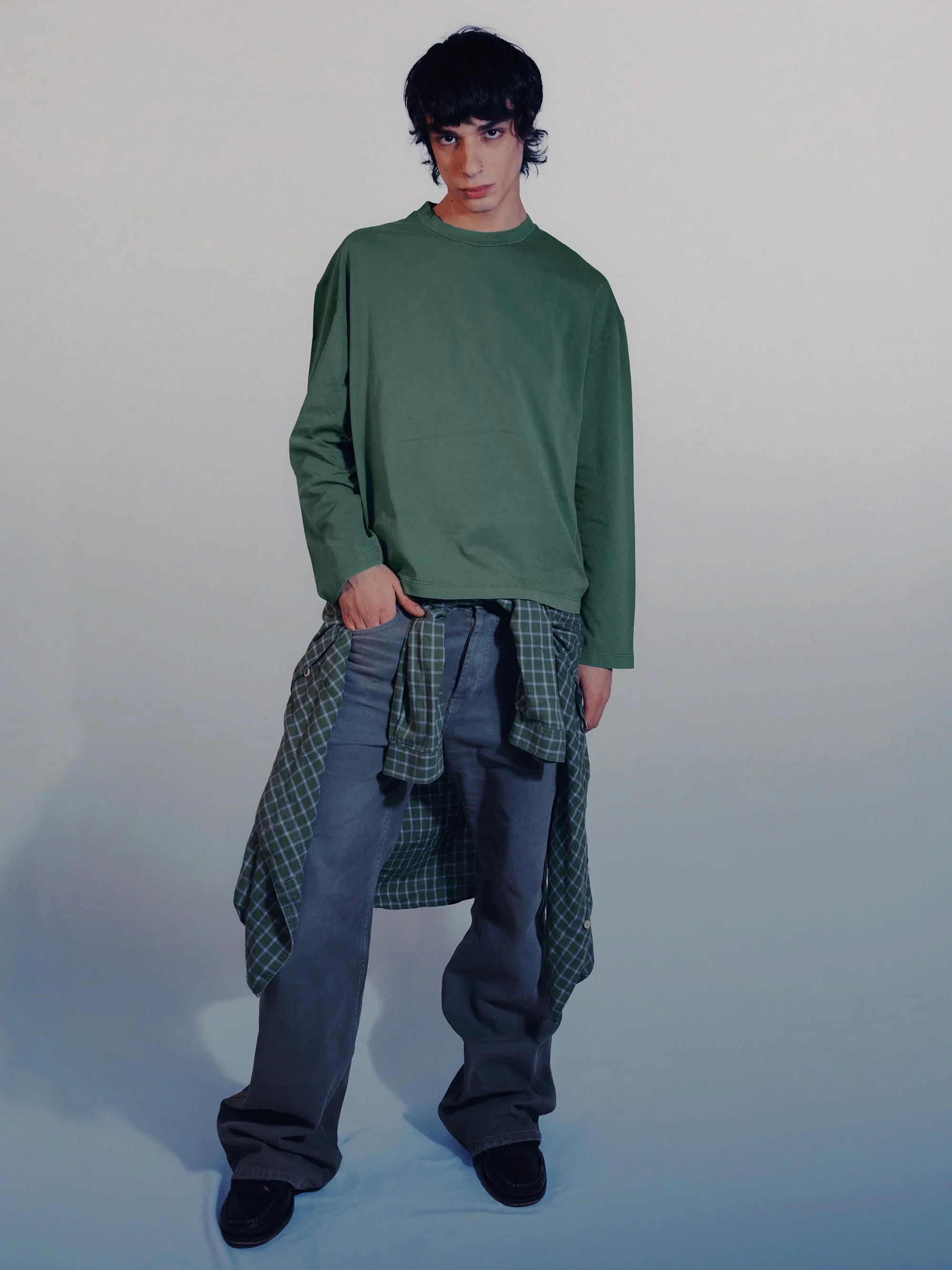 A young man with black curly hair wearing a loose green long-sleeve shirt, gray pants, and black shoes. He has a green checkered shirt tied around his waist and poses with one hand in his pocket against a plain light background.