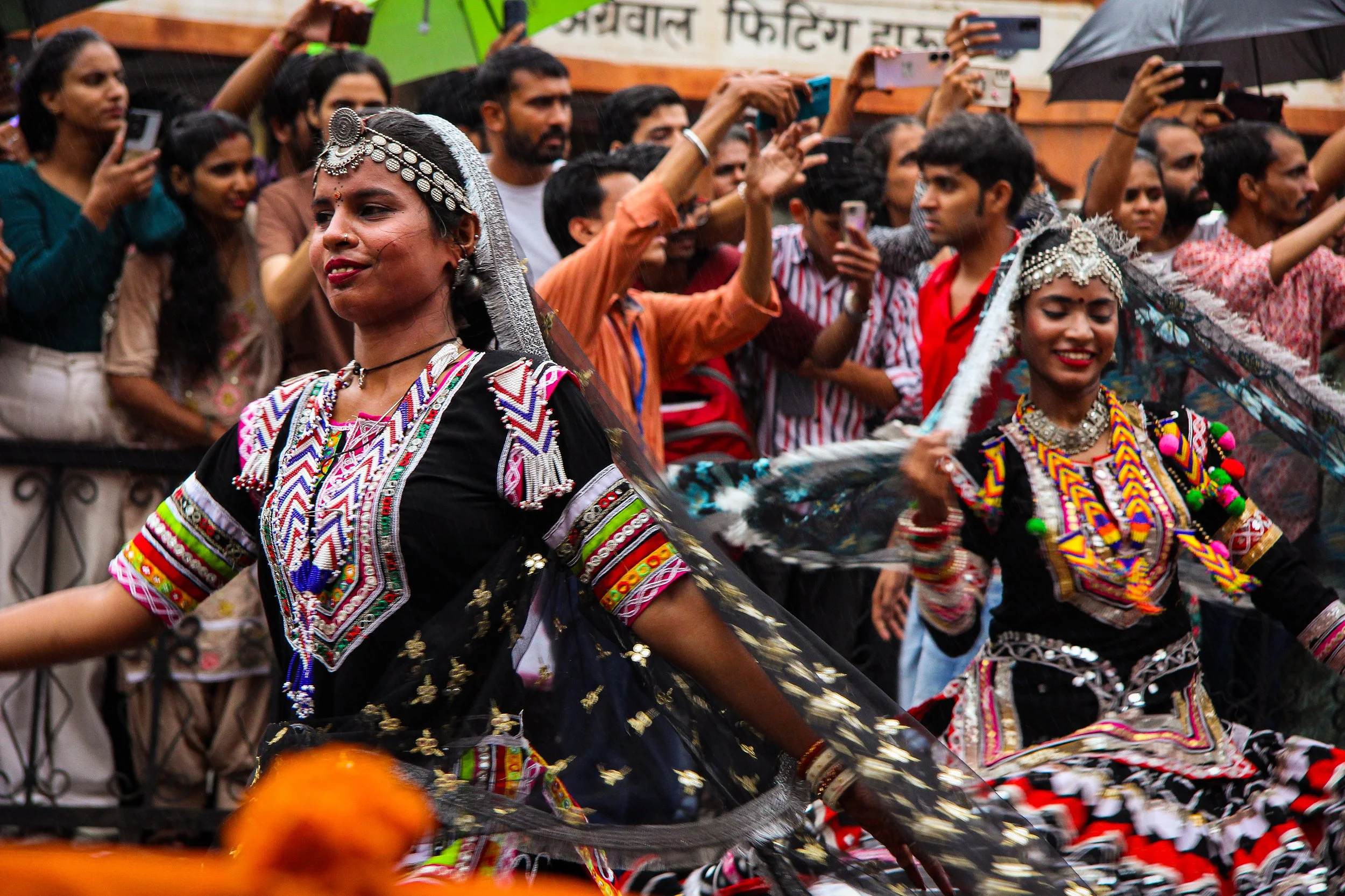 Women dressed in traditional, colorful Indian attire performing a dance on a street during a cultural celebration or festival, with a crowd of spectators taking photos and watching.