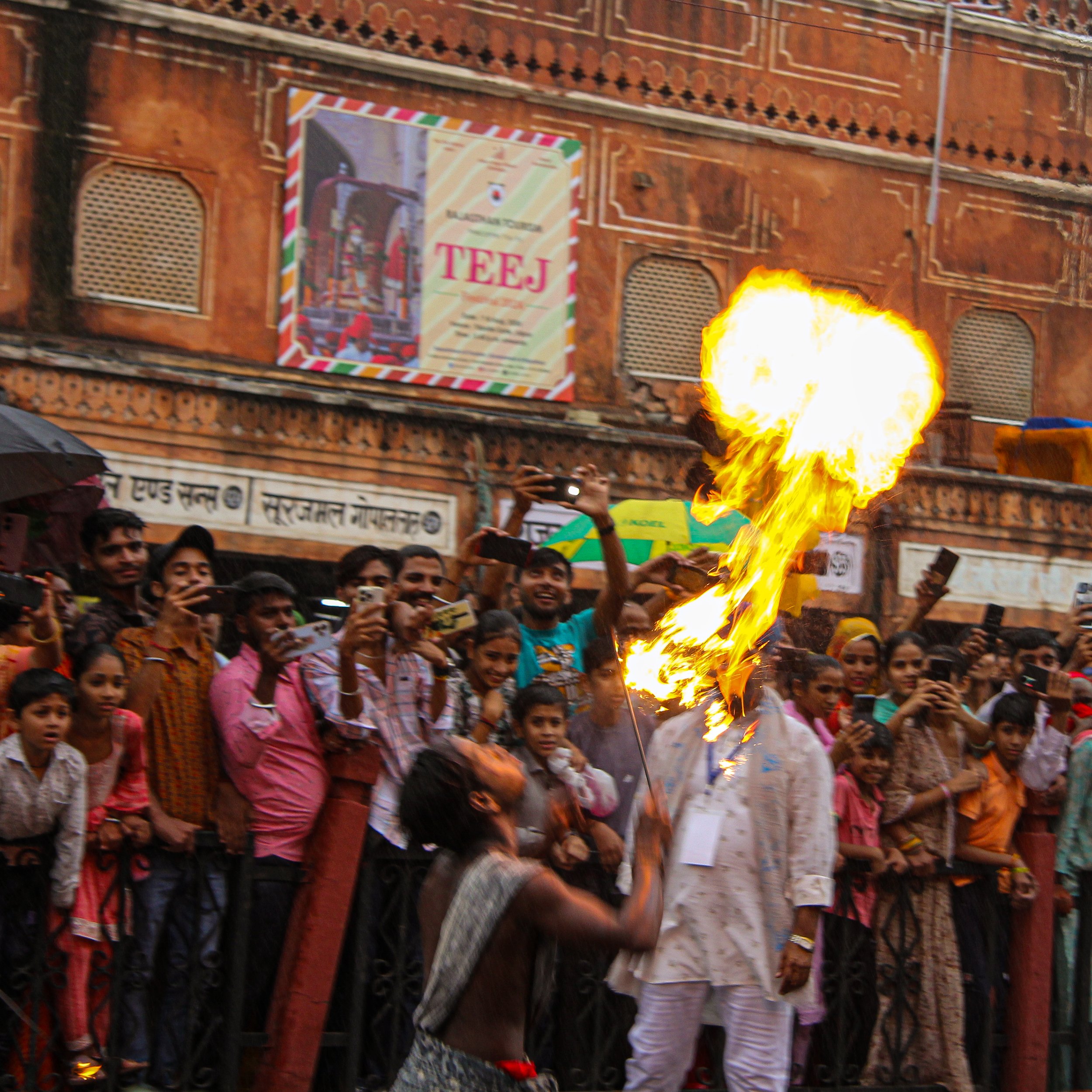 A street performer in traditional attire performs a fire-breathing act in front of a crowd of onlookers, with many taking photos. The background features an old brick building with signs and a large poster.