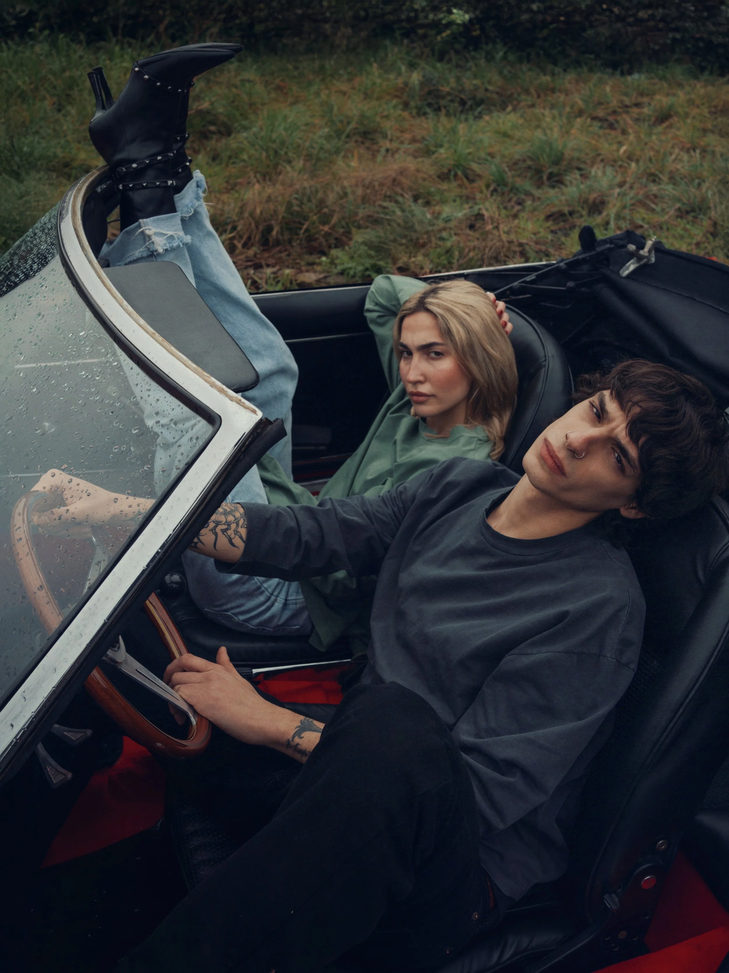 A young man and woman sitting in a vintage convertible car parked on grass, with the woman reclining and the man holding the steering wheel, both looking at the camera.
