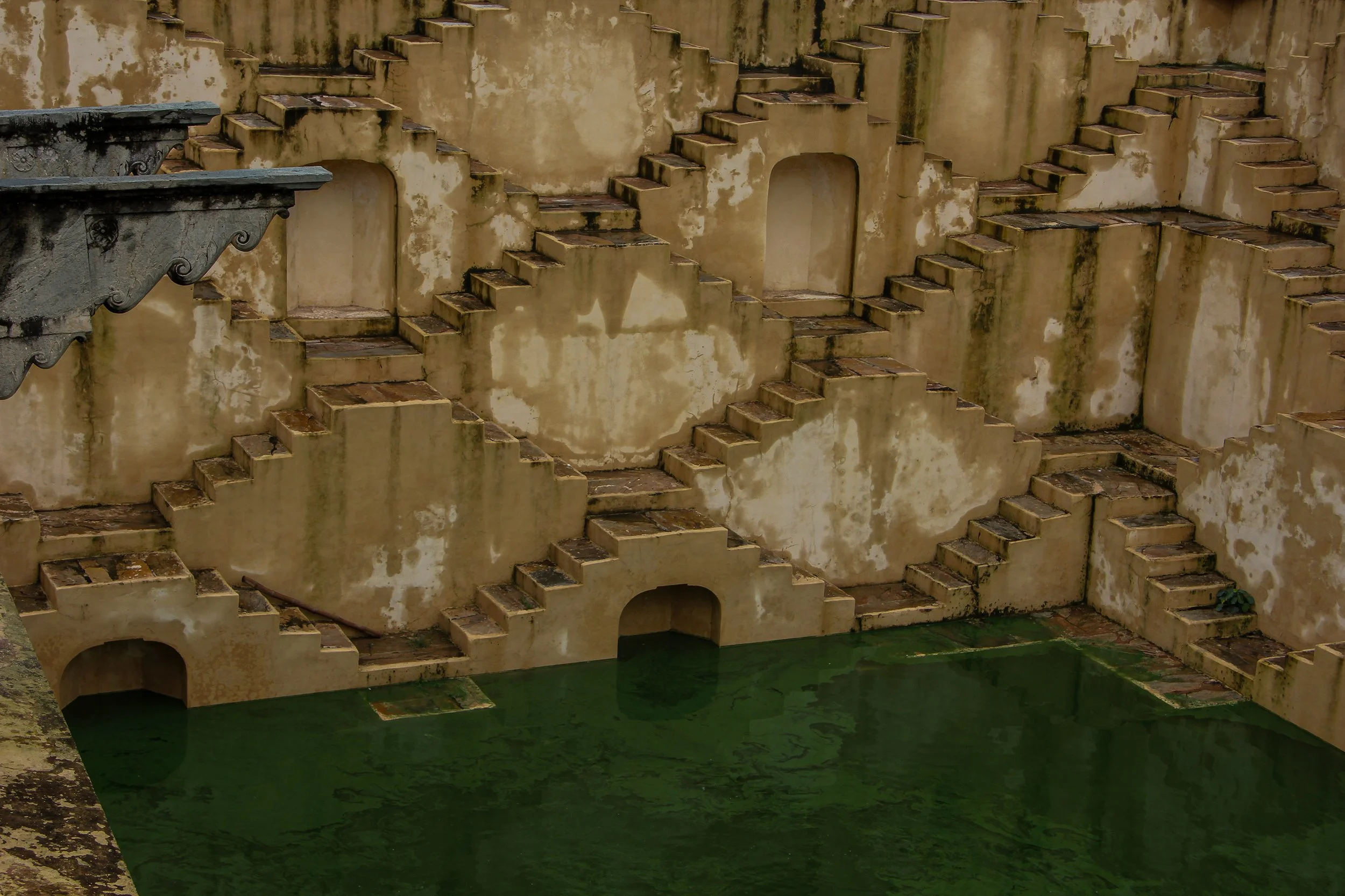 Old, weathered step and wall structures with small archways, next to a pool of still, green water.