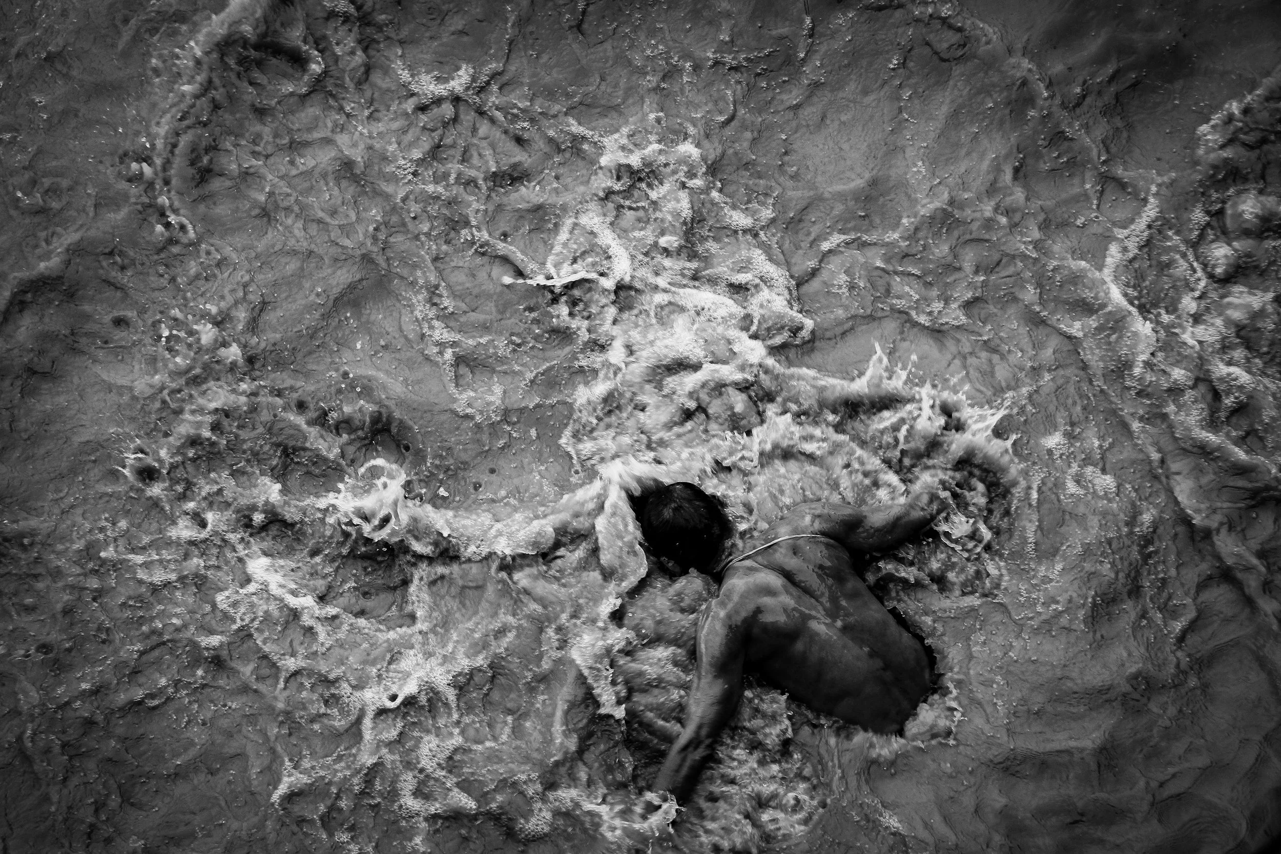 A person with short hair, wearing a necklace, is crouched in muddy water, creating splashes as they splash the water around.