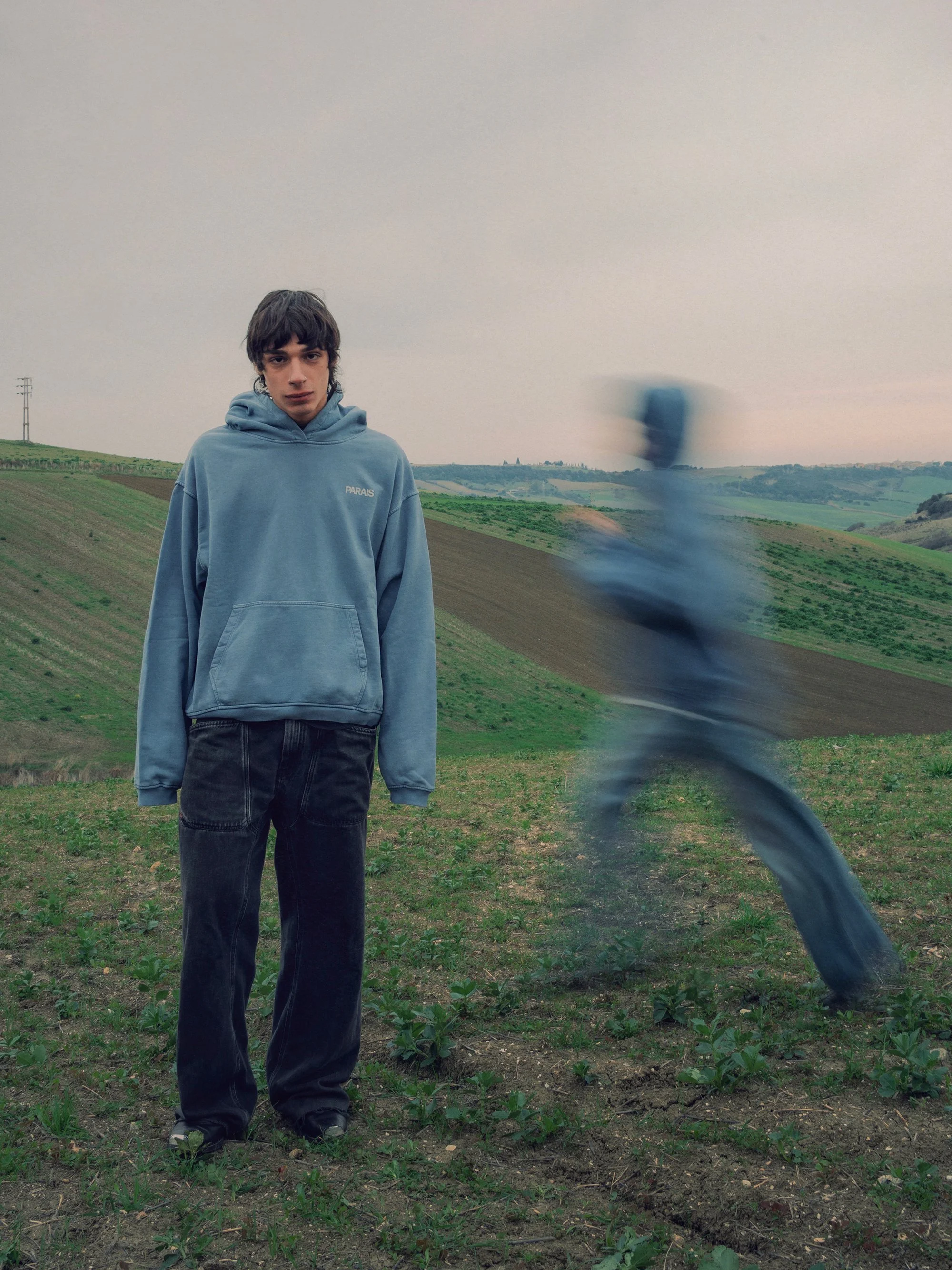 A young man in a blue hoodie standing in a rural field with rolling hills in the background. A blurred figure in motion appears to be jumping or running nearby.