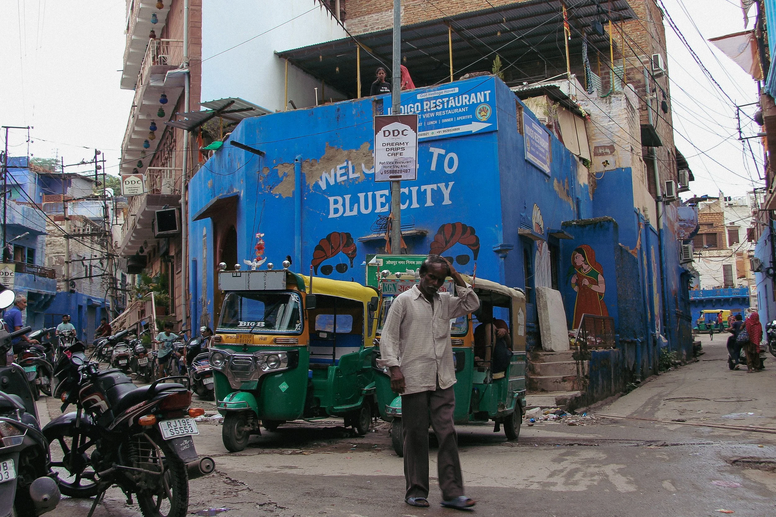 Street scene in an urban area with a blue building that has a Hello Welcome to Blue City sign, a man walking, motorcycles, auto rickshaws, and pedestrians. The building has murals, signs, and staircases.