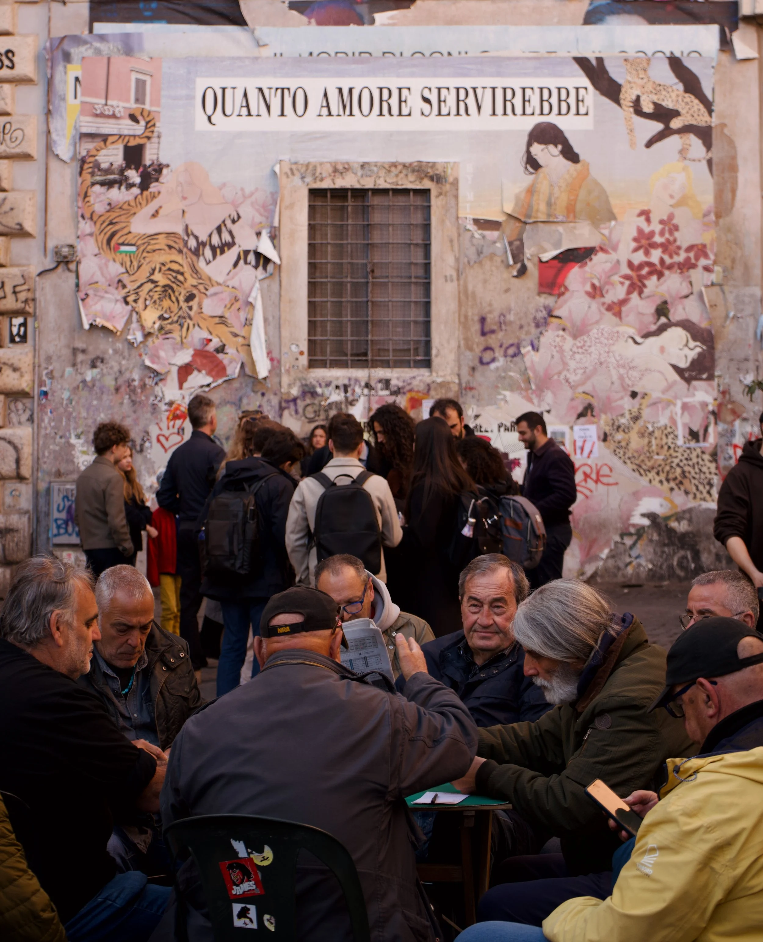 A group of people sitting and standing outdoors in front of a wall with colorful, torn posters and graffiti, including a large banner with the Italian phrase 'QUANTO AMORE SERVIREBBE'.