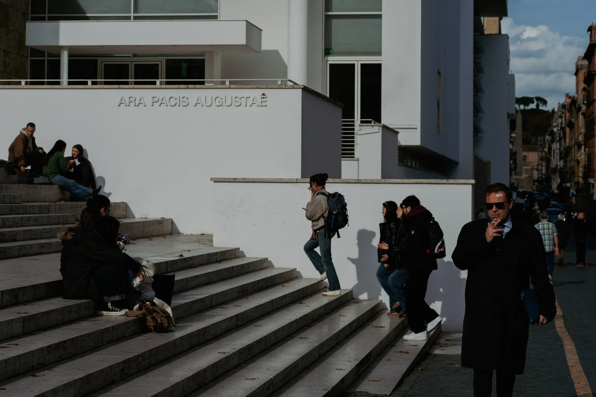 People walking up and sitting on stone steps outside a modern white building with the words 'Ara Pacis Augustae' on the wall.