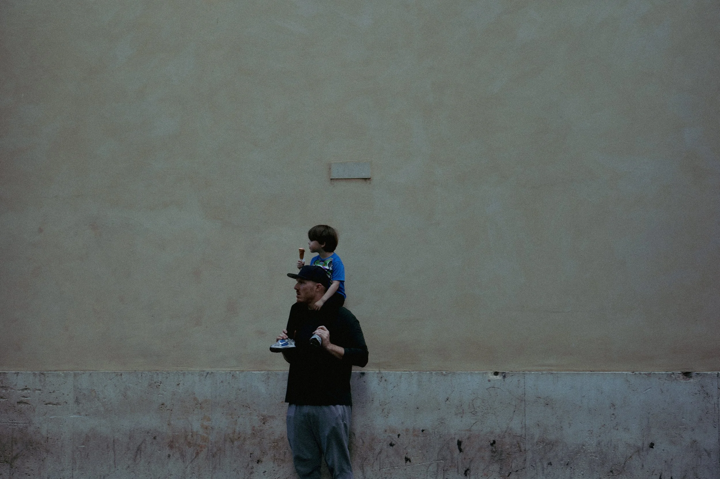 A man carrying a young boy on his shoulders, both facing left. The boy is wearing a blue shirt and holding an ice cream cone, while the man is dressed in dark clothing and a cap. They are standing against a large, plain beige wall with a small rectan