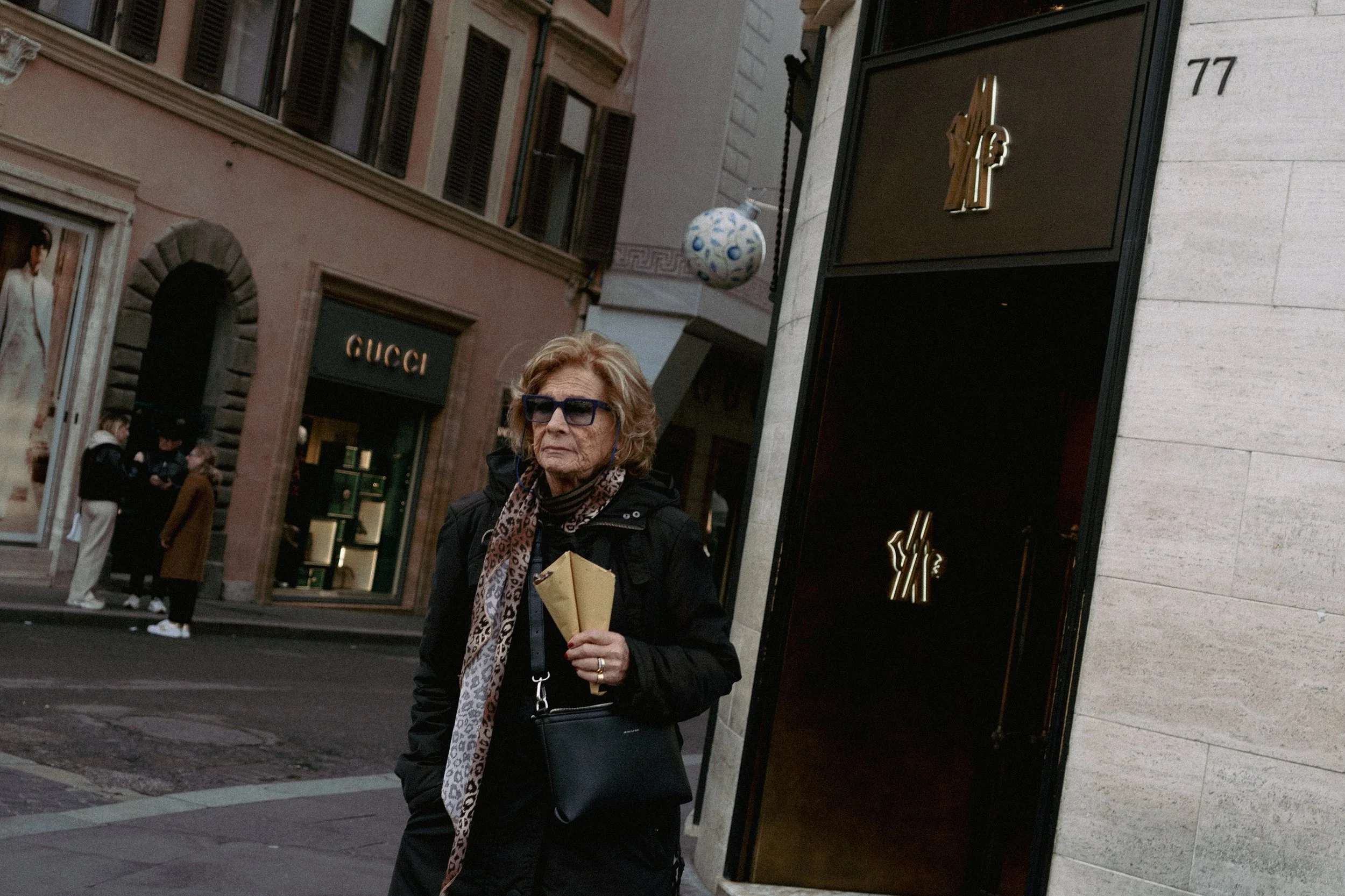 An older woman in black clothing, sunglasses, and a leopard print scarf holding two paper bags, walking outdoors in an urban area near a Gucci store.