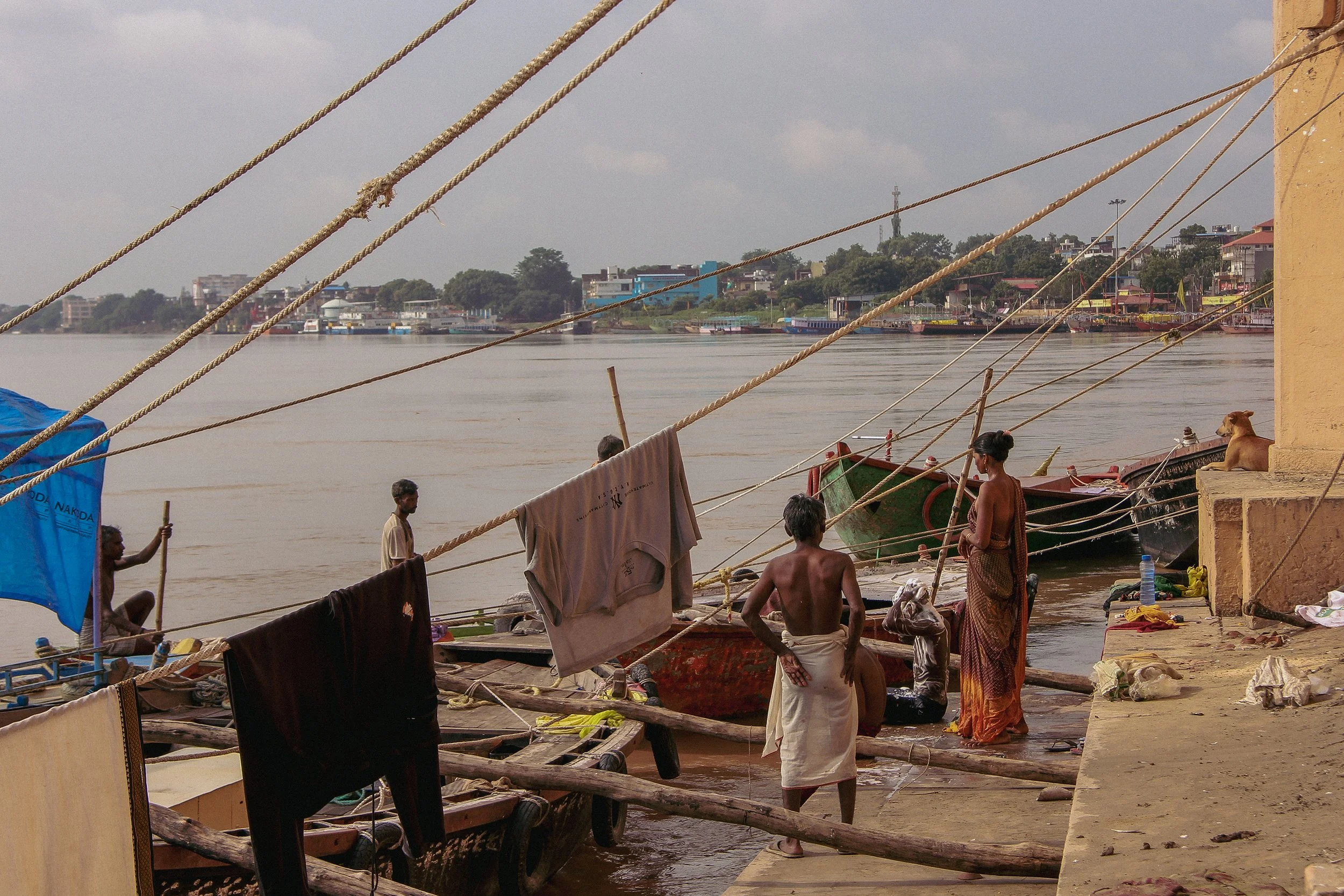 People by the river on a dock, with boats and buildings in the background.