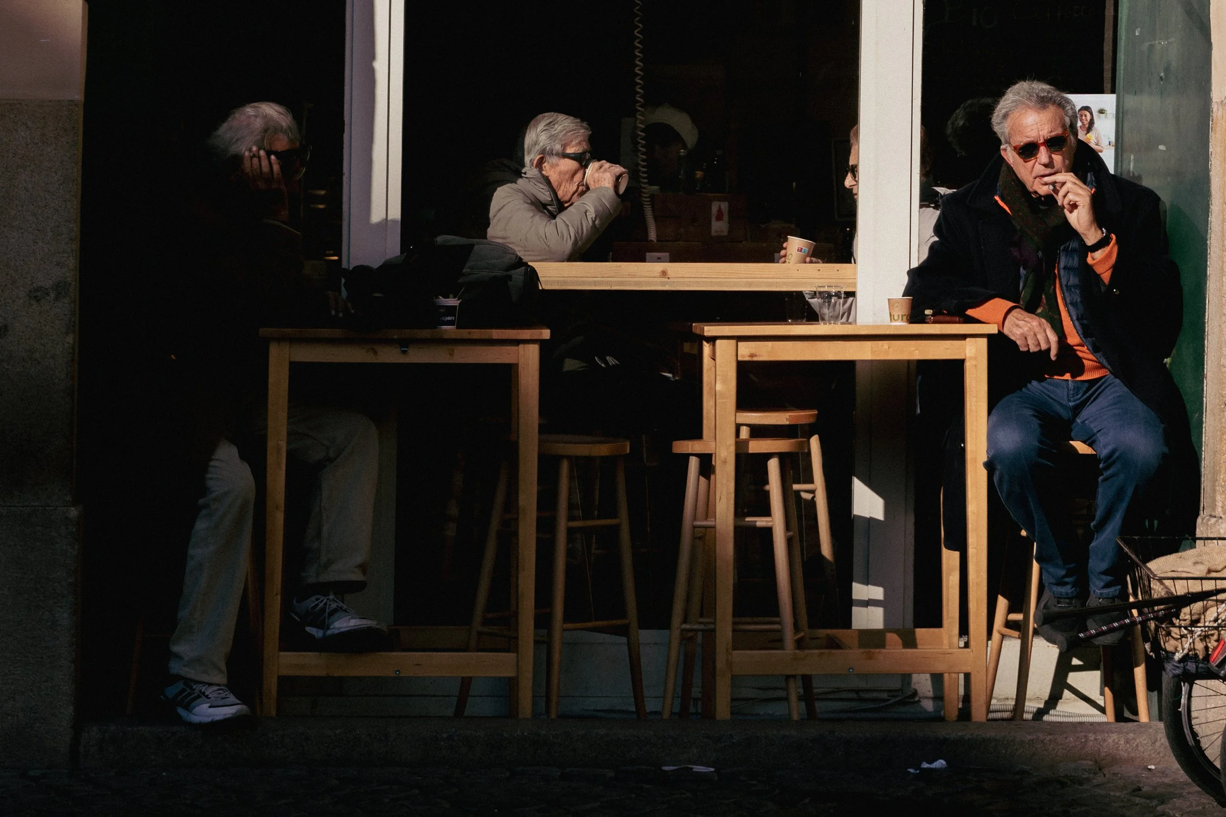 Four people sitting outside a cafe in the evening, with two people near the window and two people seated in front of it. One person is drinking from a cup, another is smoking, and others are sitting at tables.