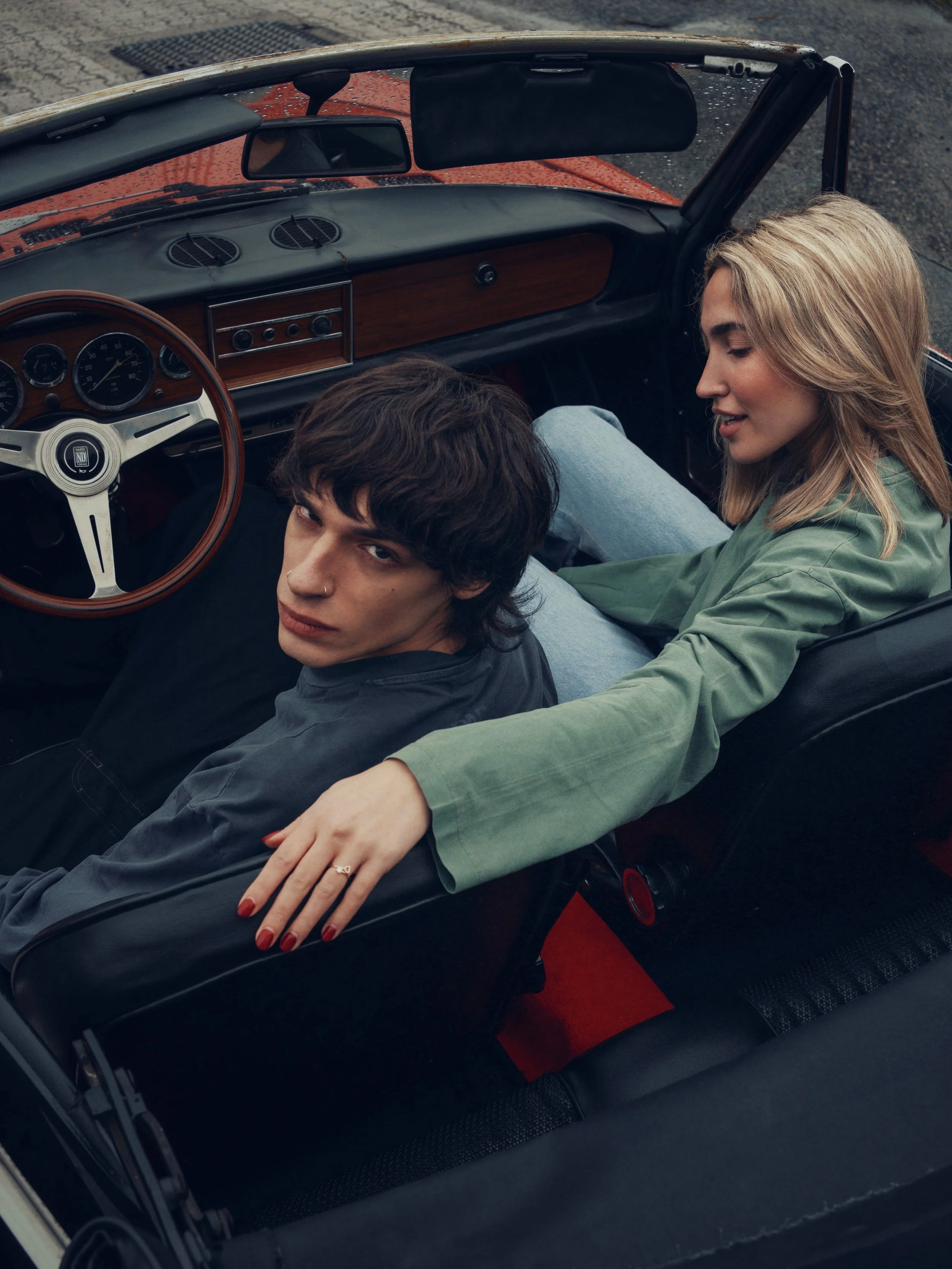 A young man and woman sitting in a vintage convertible car, with the woman sitting on the back seat, leaning over the front passenger seat, and the man in the driver's seat, looking up at the camera.