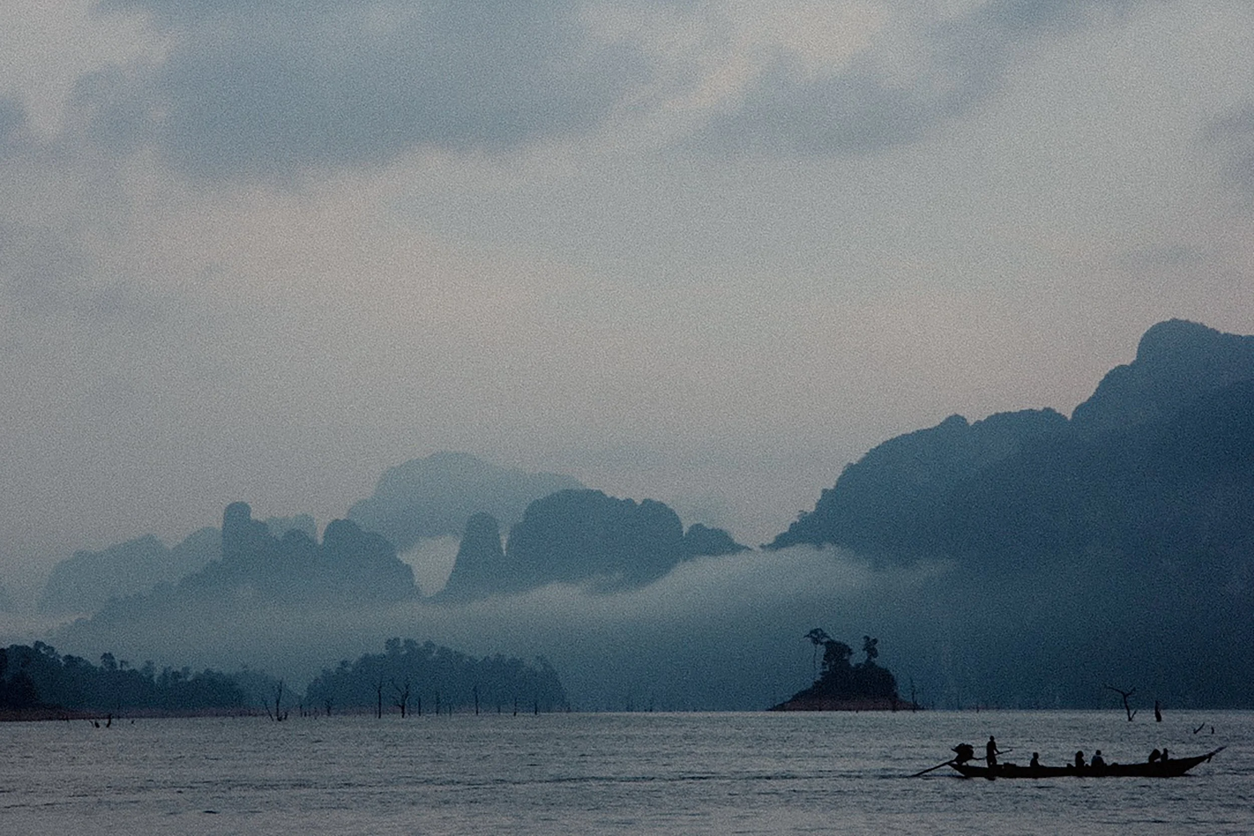 A serene body of water with a small boat carrying people, set against a backdrop of misty mountains and overcast sky.