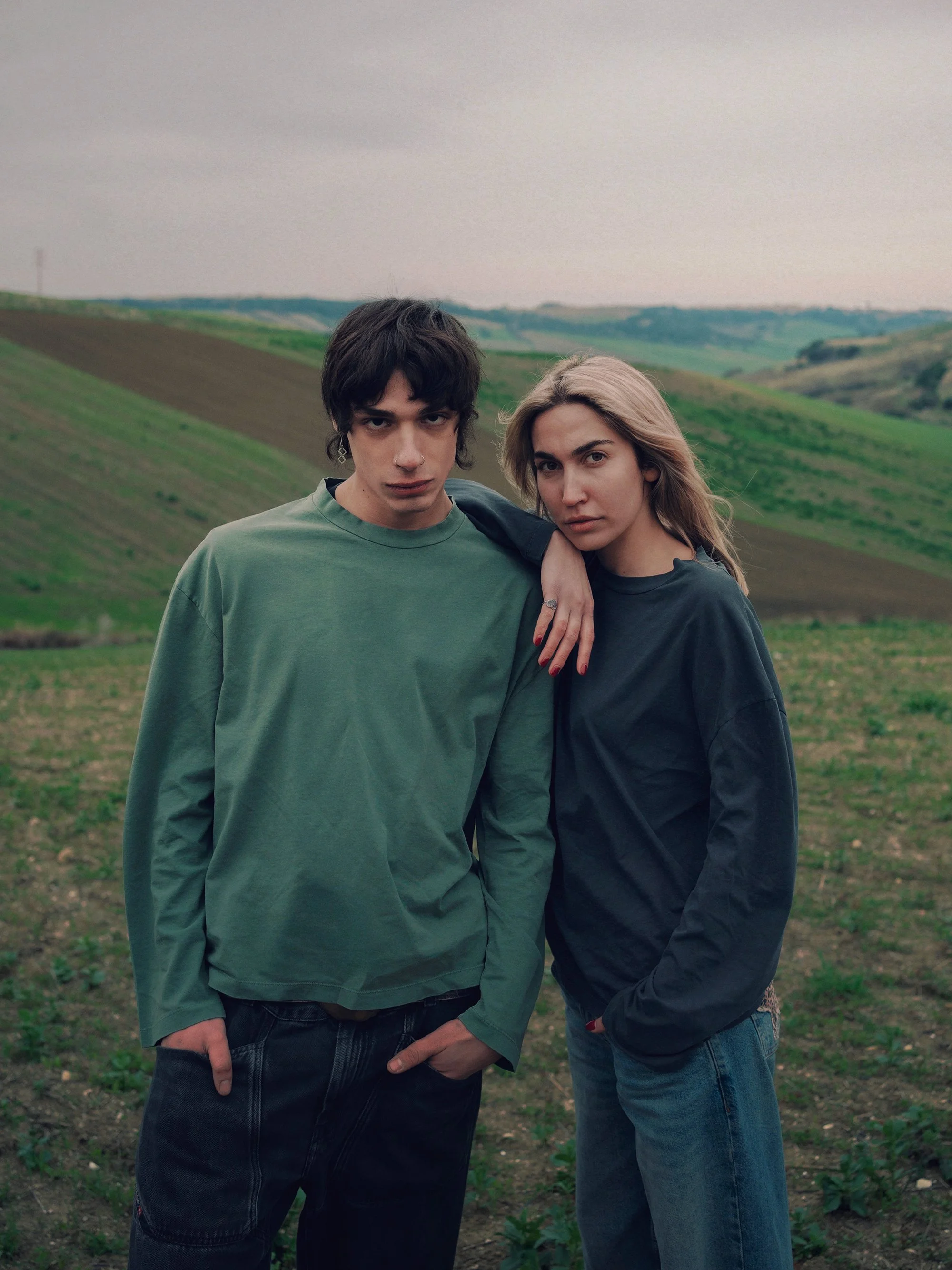 A young man and woman standing closely together outdoors in a hilly, green landscape with overcast sky.
