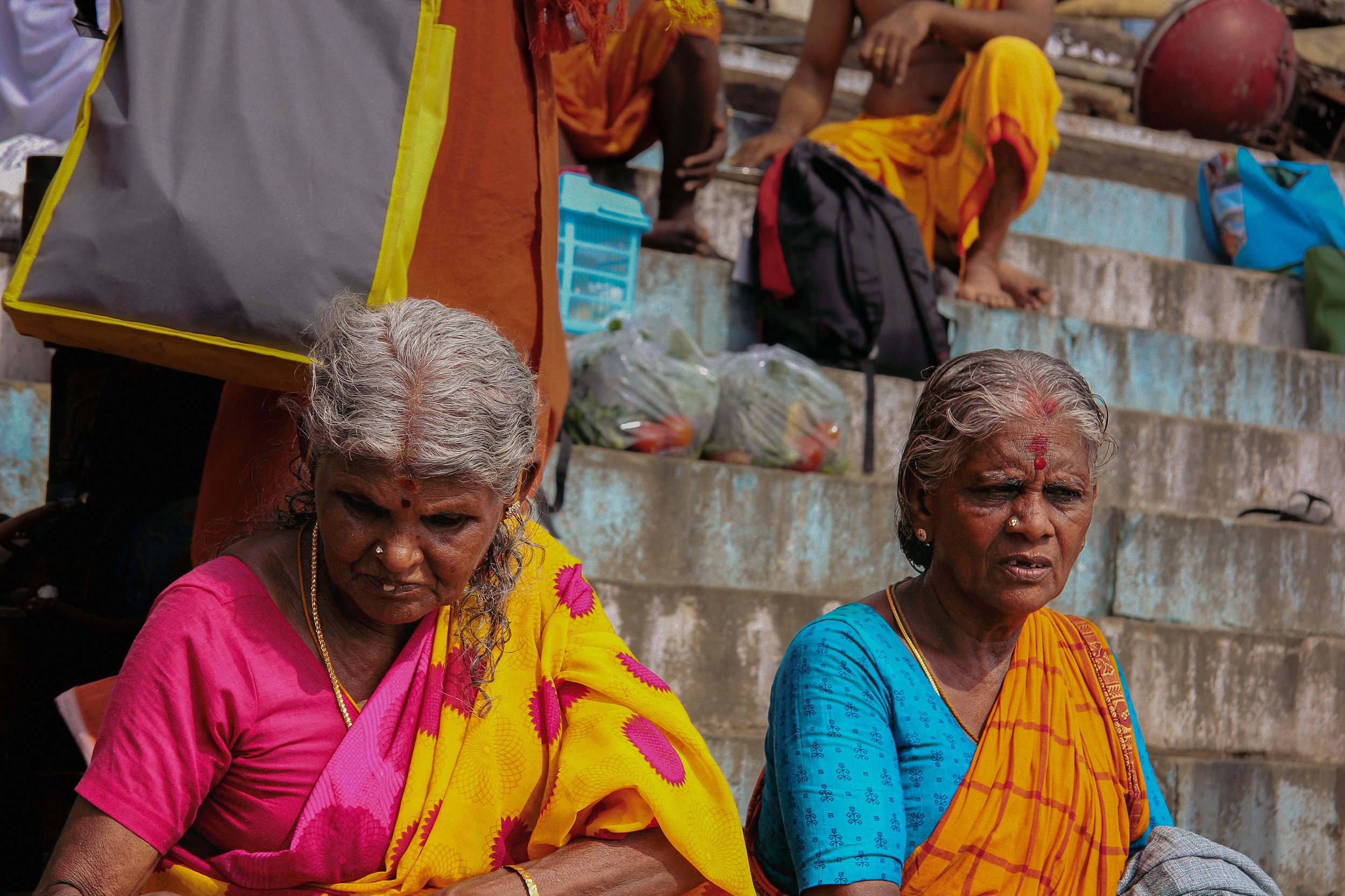 Two elderly women sitting on stone steps, wearing traditional colorful Indian sarees, with bags and backpacks around them. One woman has silver hair and the other has grey hair, both with reddish marks on their foreheads.