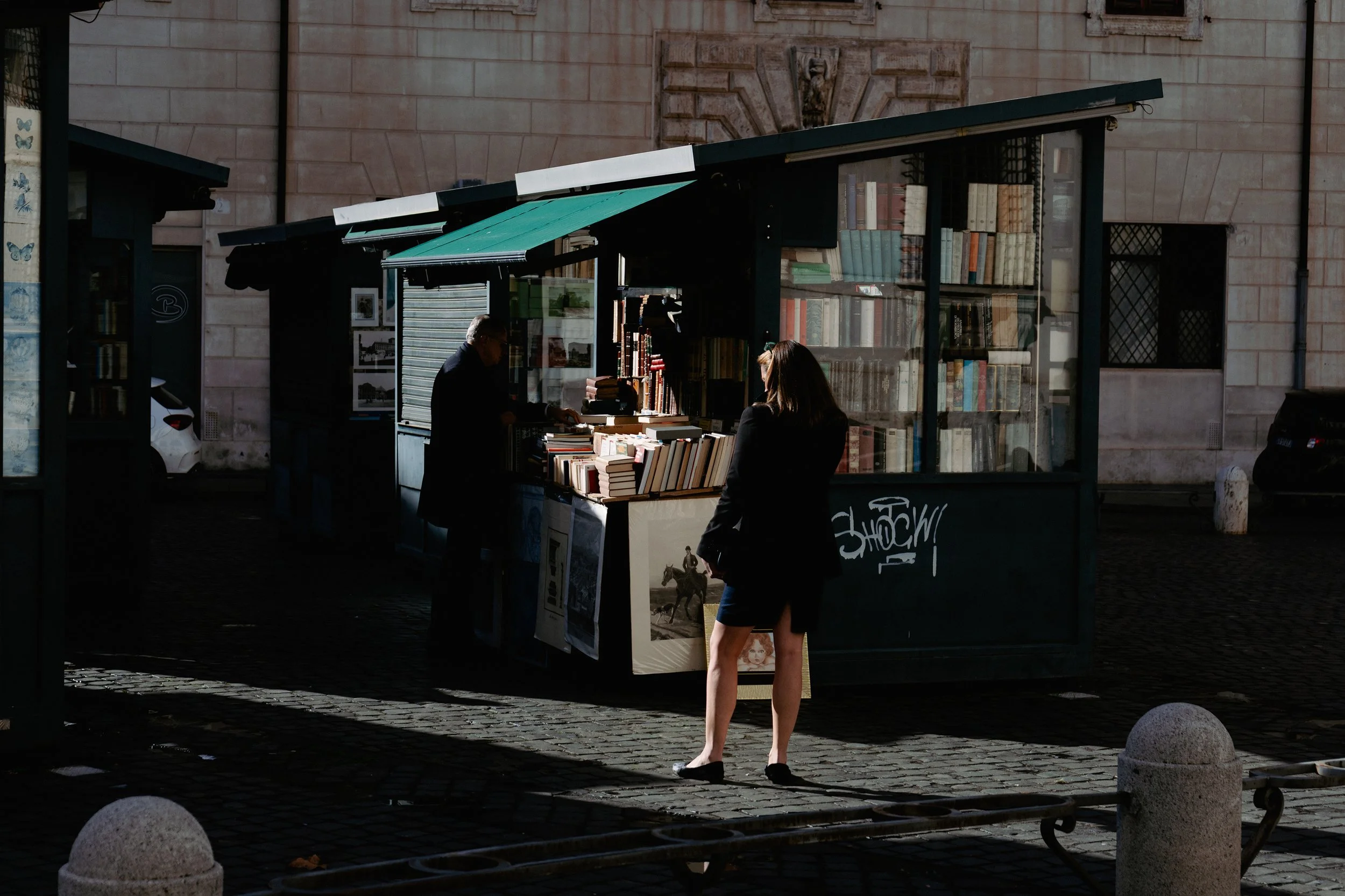 A woman in a black blazer and skirt is standing in front of an outdoor bookstore, browsing books while a man behind the stall arranges books on display. The bookstore has a small glass wall and green awnings, with books stacked on tables and inside t