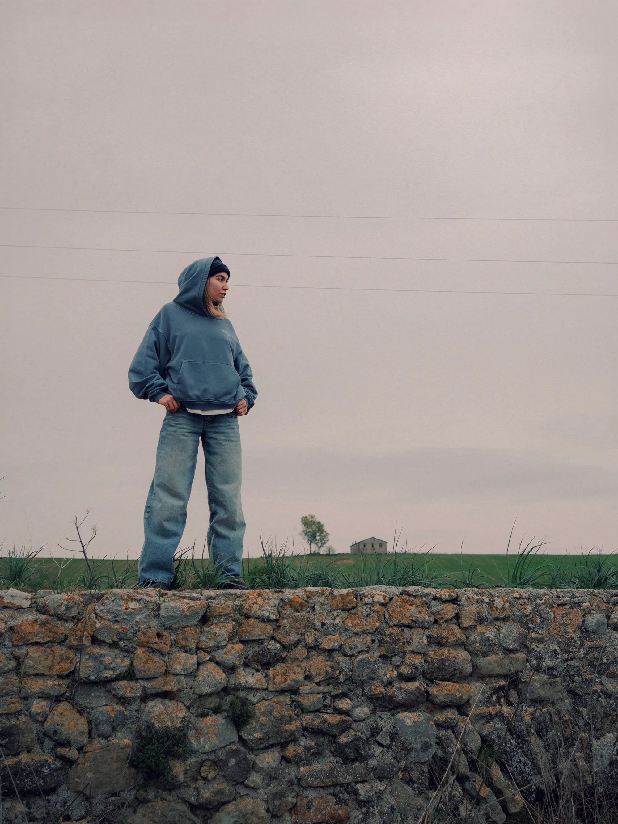 Person standing outdoors on a stone wall wearing a blue hoodie and jeans, looking to the side on an overcast day.