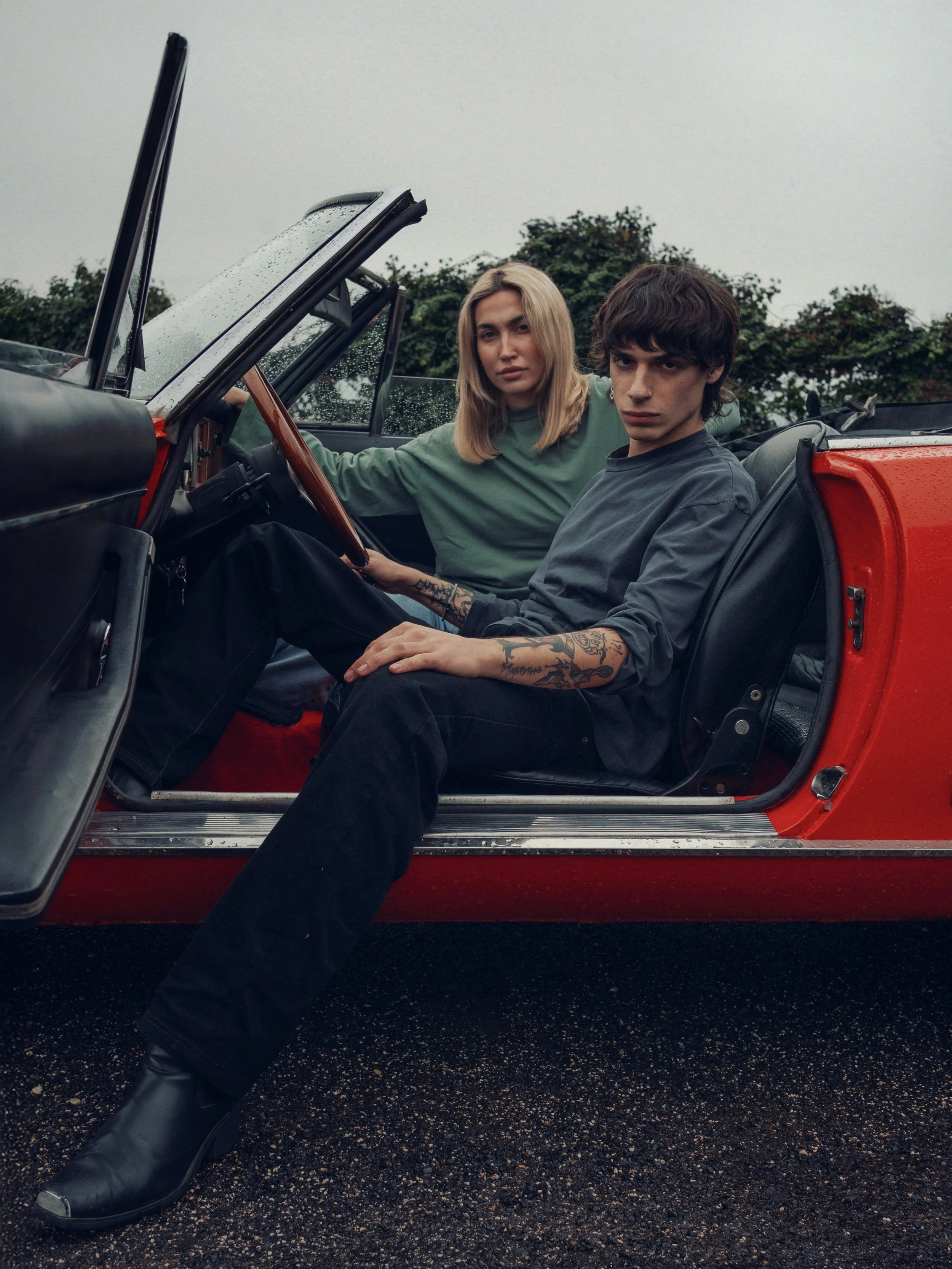 A young woman and man sitting in an open-top red vintage car on a rainy day, with the woman driving and the man sitting in the passenger seat. They appear serious, with dark clothing and tattoos visible on the man.