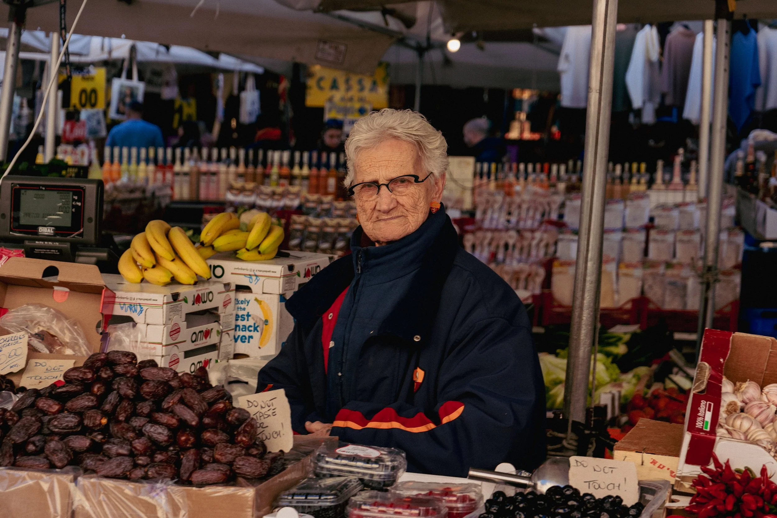 An elderly woman with glasses and earrings stands behind a produce stall at an outdoor market, with bananas, dates, garlic, and other produce on display.