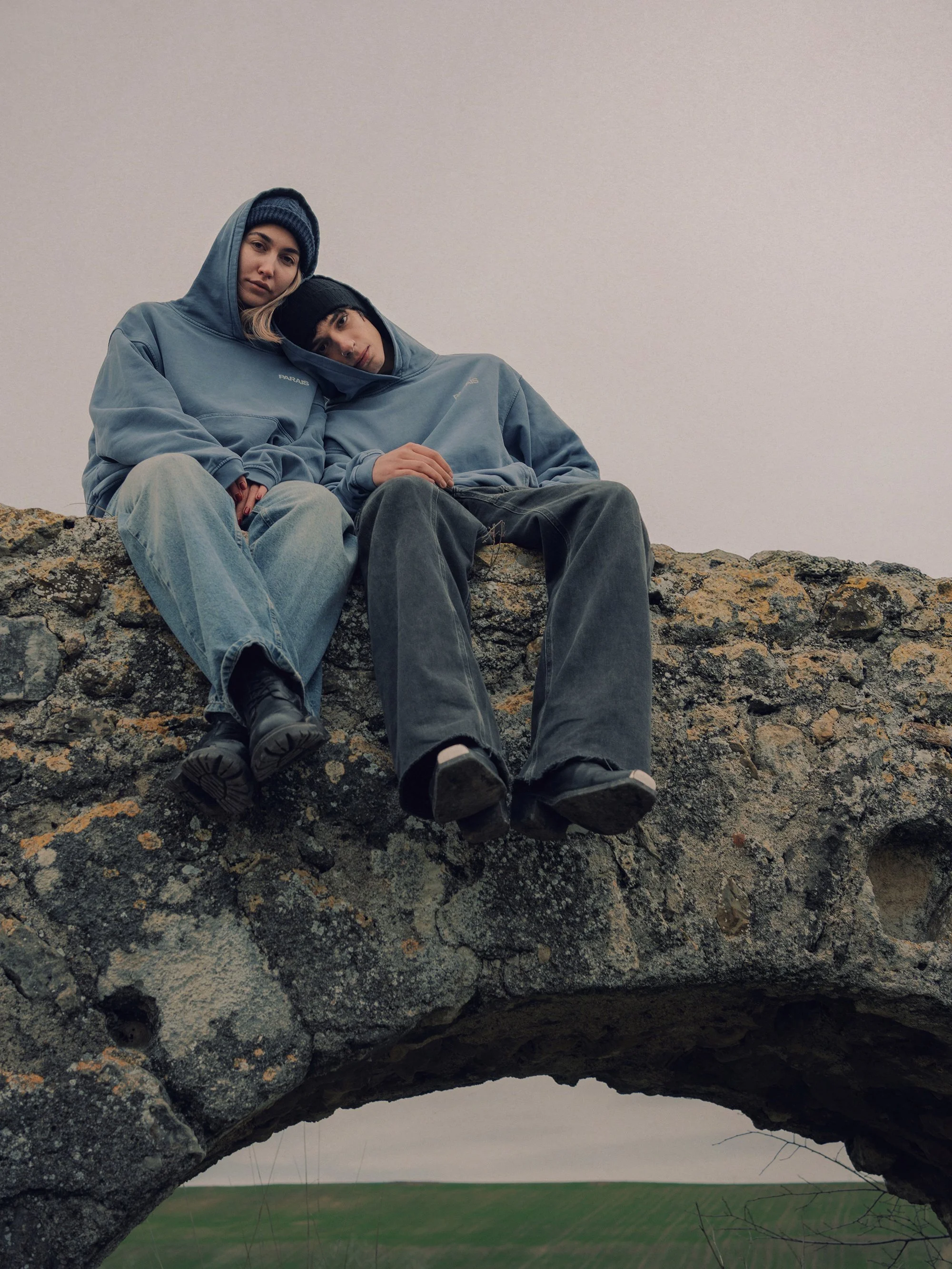 Two people wearing blue hoodies and beanies sitting on a stone arch bridge, looking at the camera with neutral expressions.