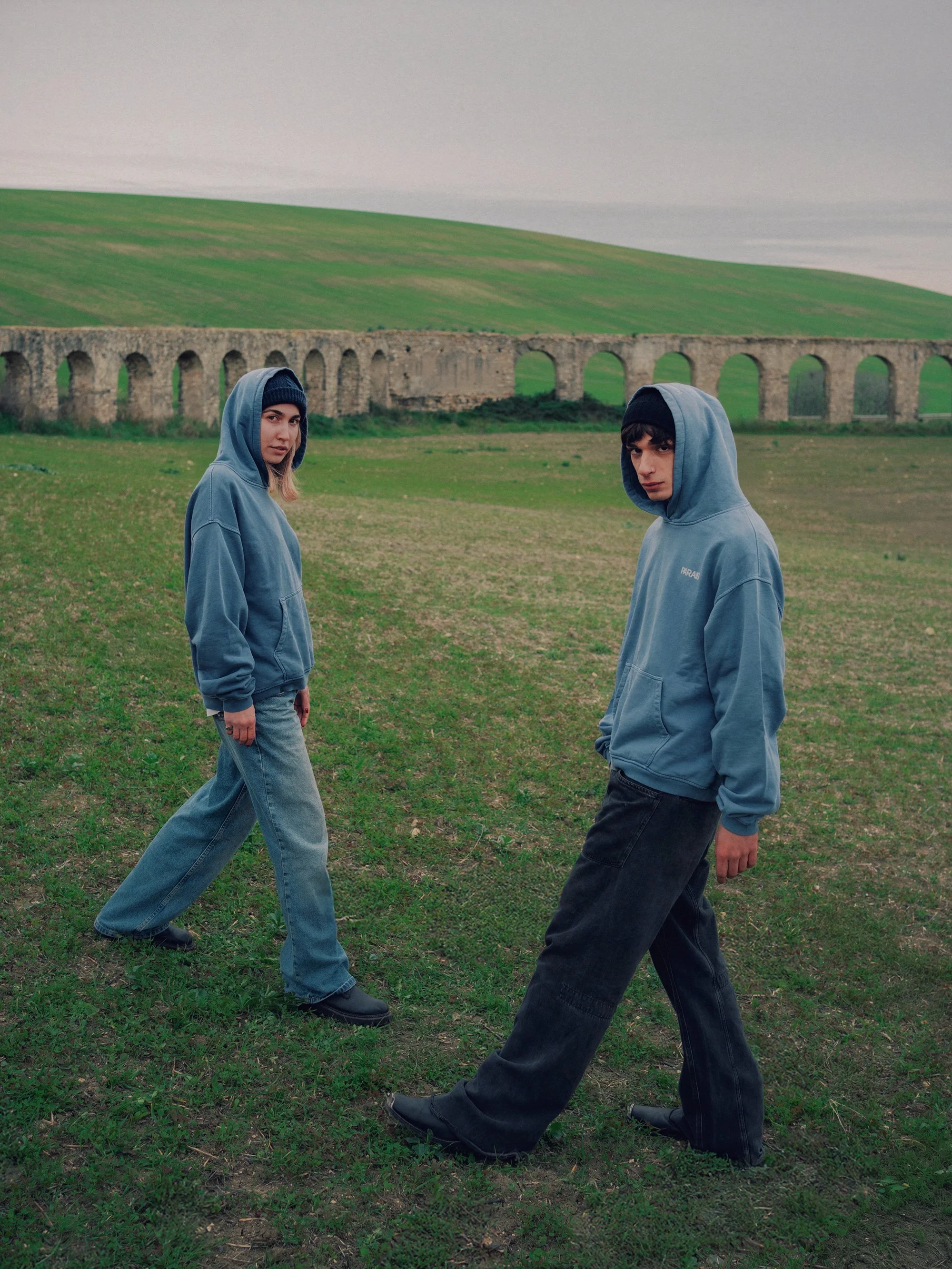 Two young people wearing blue hoodies and jeans walking on grass with an ancient stone aqueduct and green rolling hills in the background.