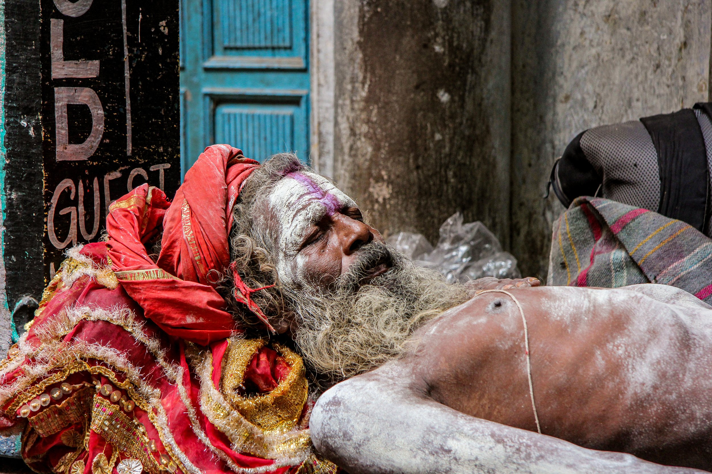 An elderly man with a long beard and traditional markings on his face, dressed in vibrant red and gold clothing, lying on the ground with his eyes closed.