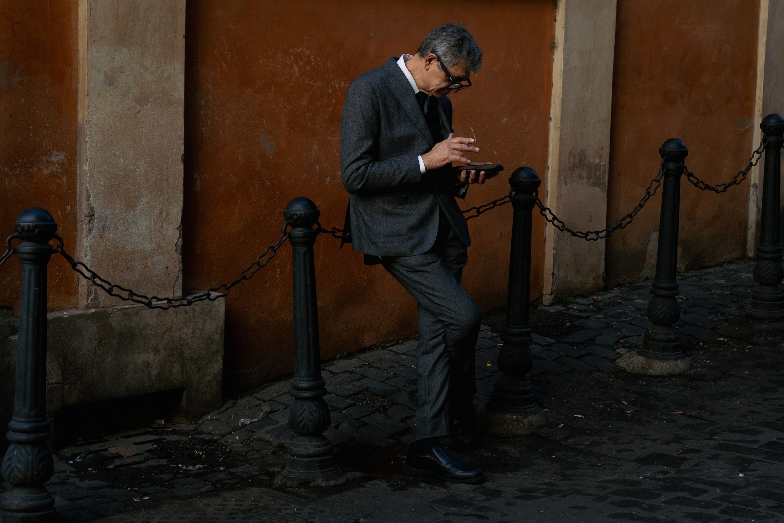 A man in a grey suit and black shoes leans against black chain-linked stanchions on a cobblestone street, looking at his phone.