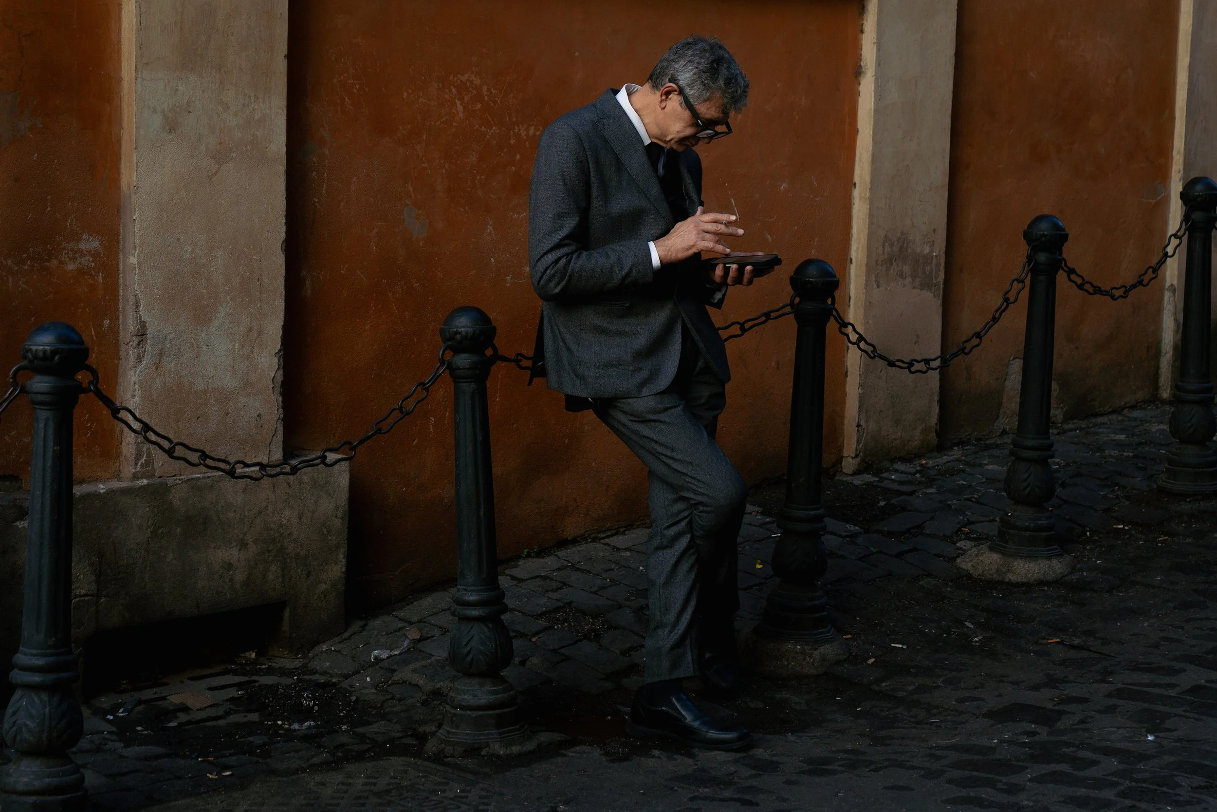 A man in a dark suit and glasses standing outdoors on a cobblestone street, leaning against a chain barrier, looking at a tablet or phone.