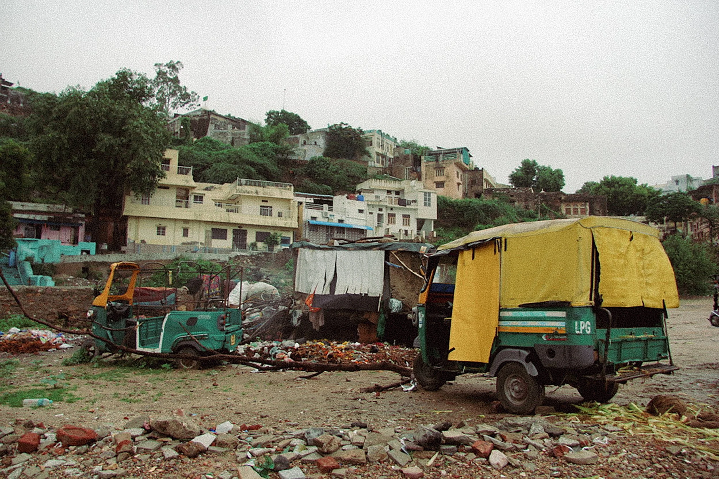 A dry, dusty open area with rubble and debris, including a small green auto rickshaw and a green LPG cargo tricycle, with a backdrop of hillside houses and scatterings of trees.