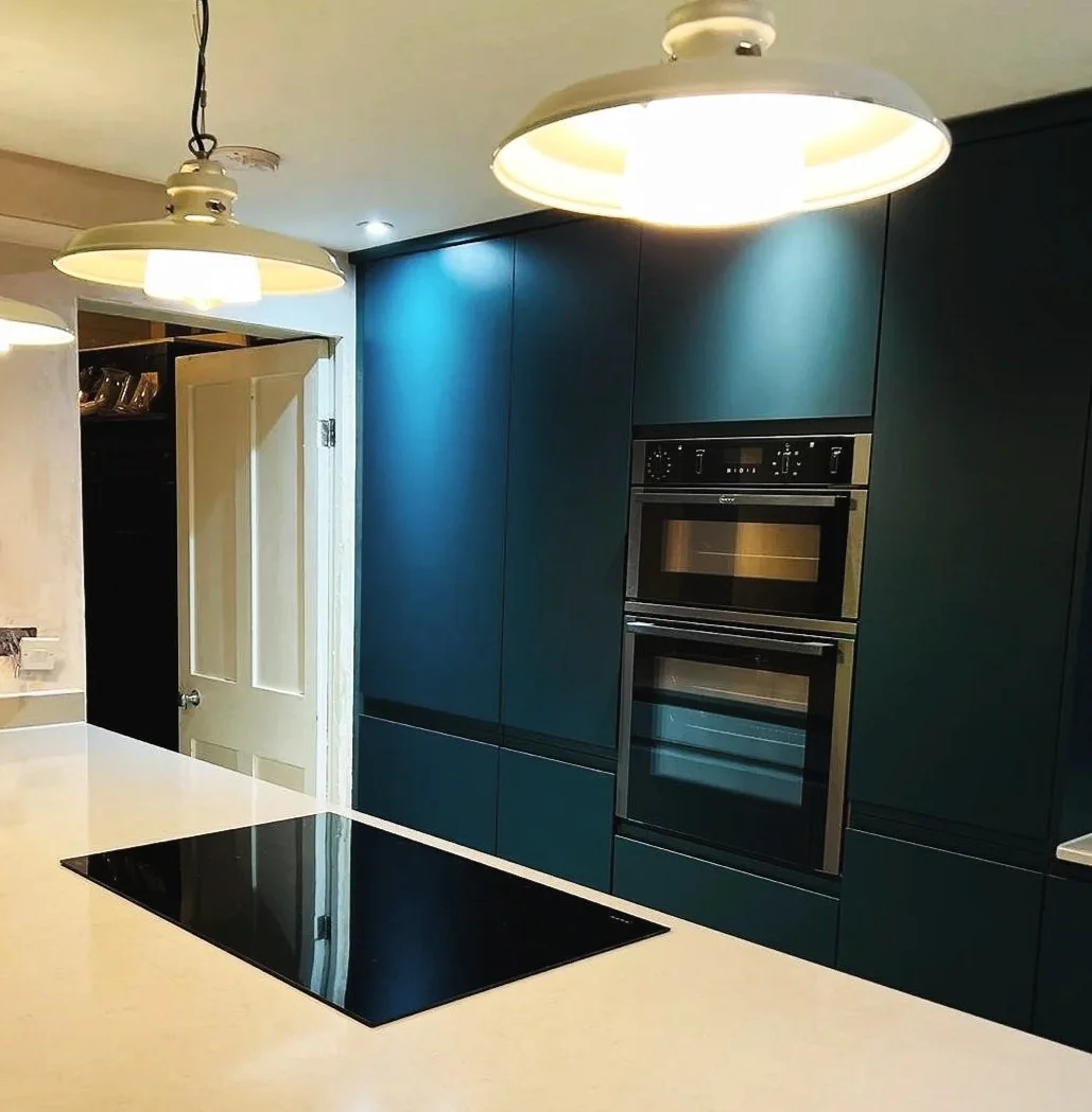 Modern kitchen with teal cabinetry, built-in oven, and black electric cooktop on cream countertop. Overhead lighting fixtures are also visible.