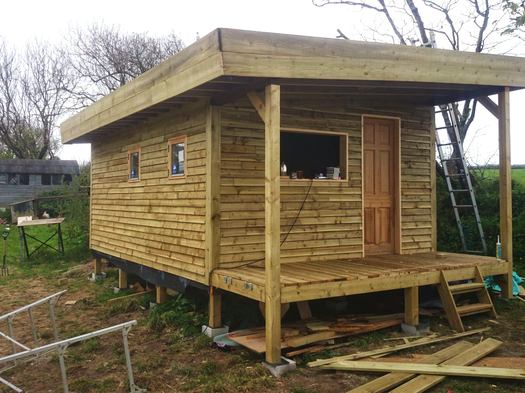 A small wooden house under construction, elevated on stilts with a porch, ladder, and two small windows.
