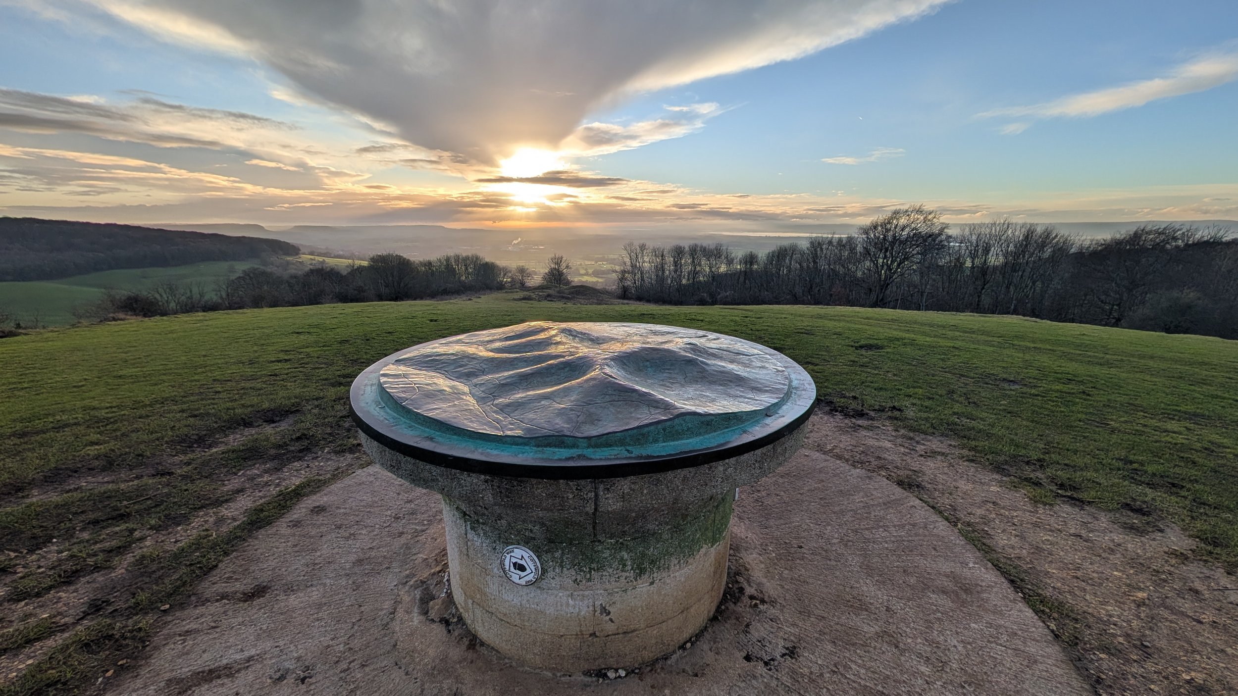 A scenic view of a sunset over a countryside landscape with a wide grassy field, trees, and rolling hills. In the foreground, there is a circular stone structure with a glass top, possibly a sundial or an elevated map, situated on a concrete area.