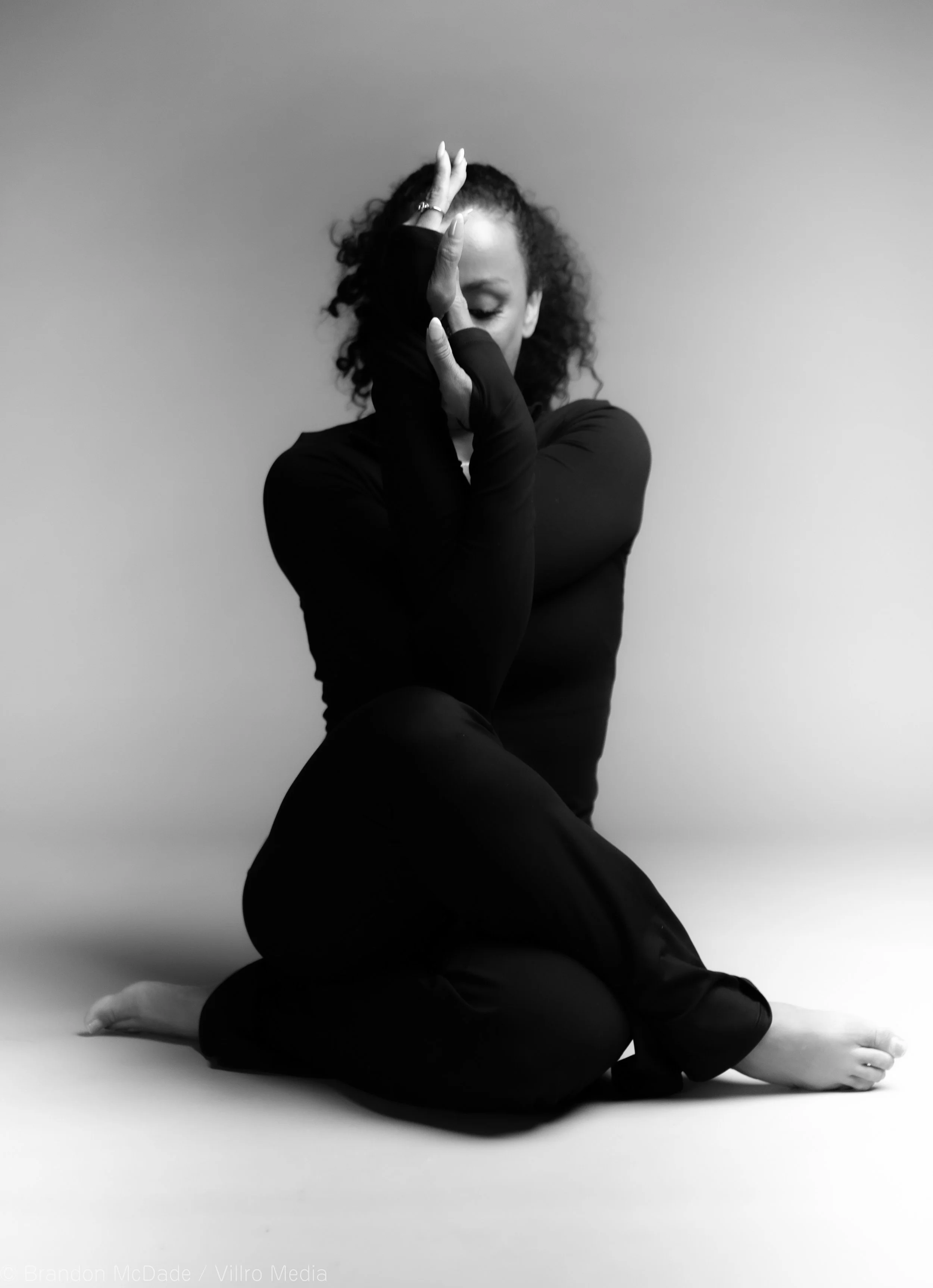 A woman with curly hair sitting on the floor, dressed in black, with her eyes closed, touching her face with her hands in a dance pose against a plain background.