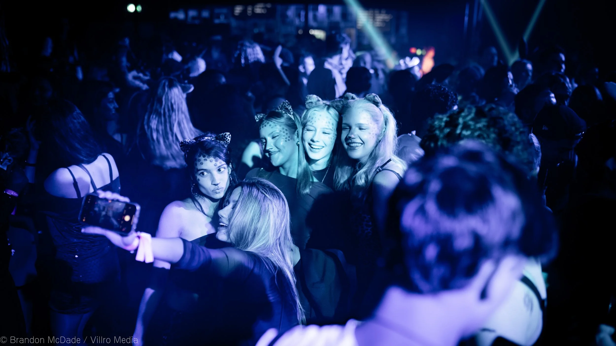 Group of young women with cat ears and face paint taking selfies and enjoying a lively dance party with colorful lights.