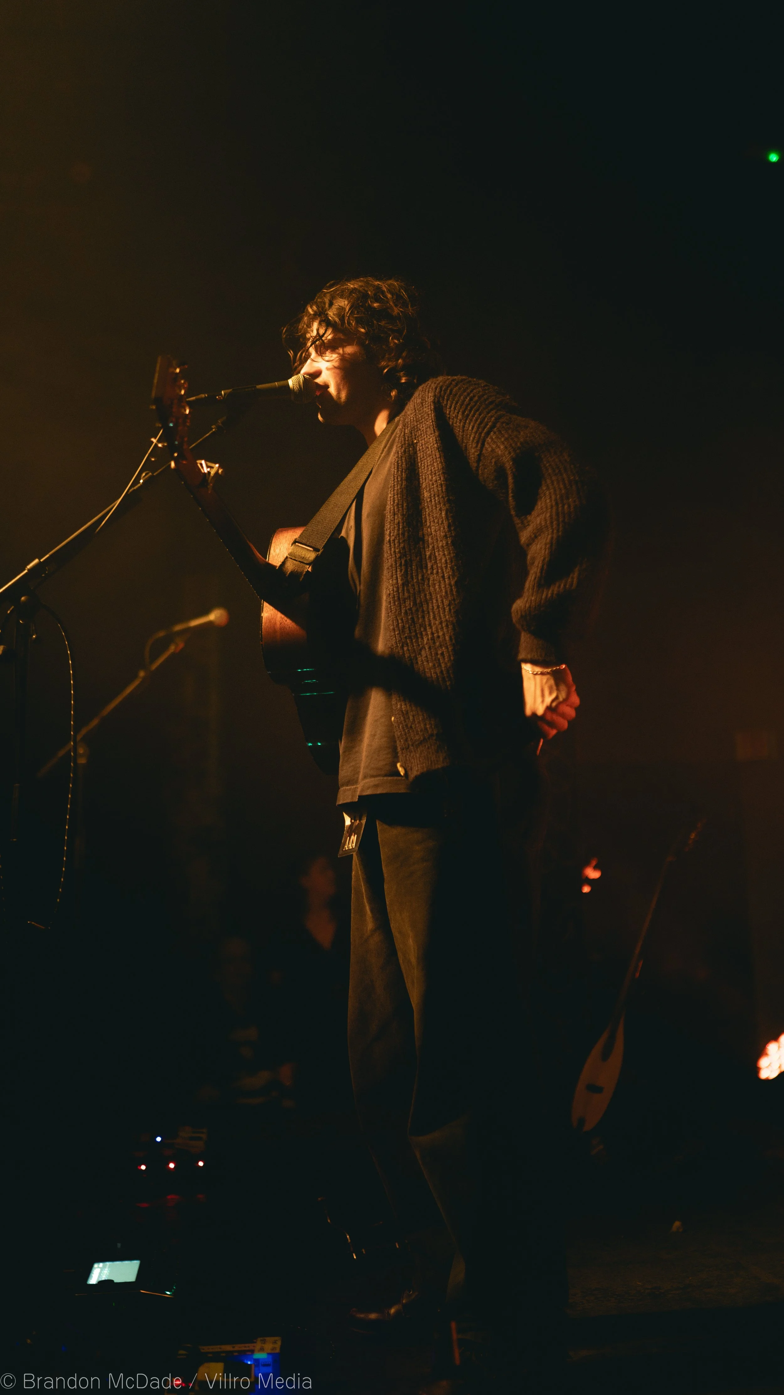 A male musician with dark, curly hair singing into a microphone while playing an acoustic guitar on a dimly lit stage.