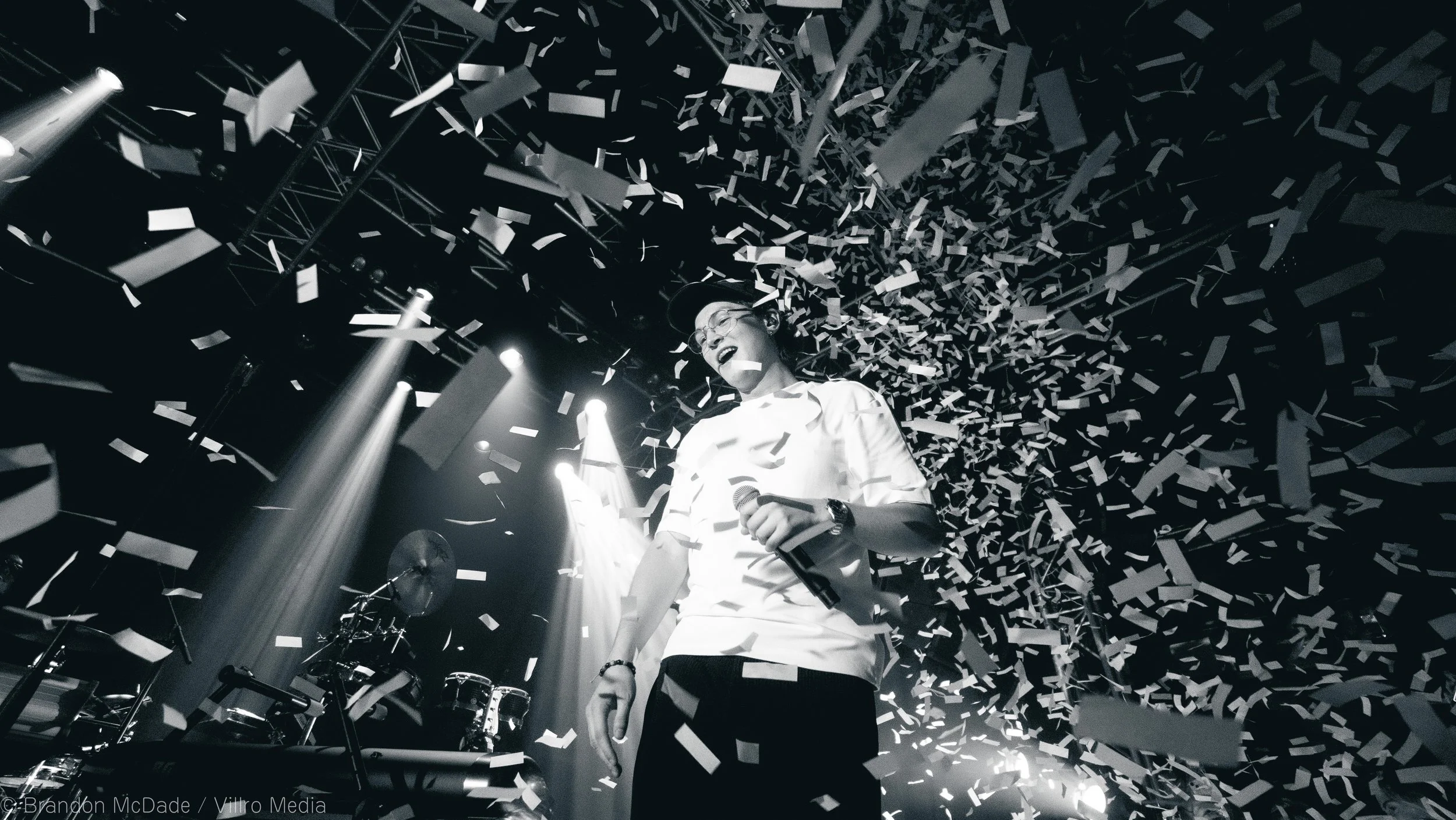 A person on stage under bright spotlights with confetti falling around, celebrating a moment of victory or excitement.
