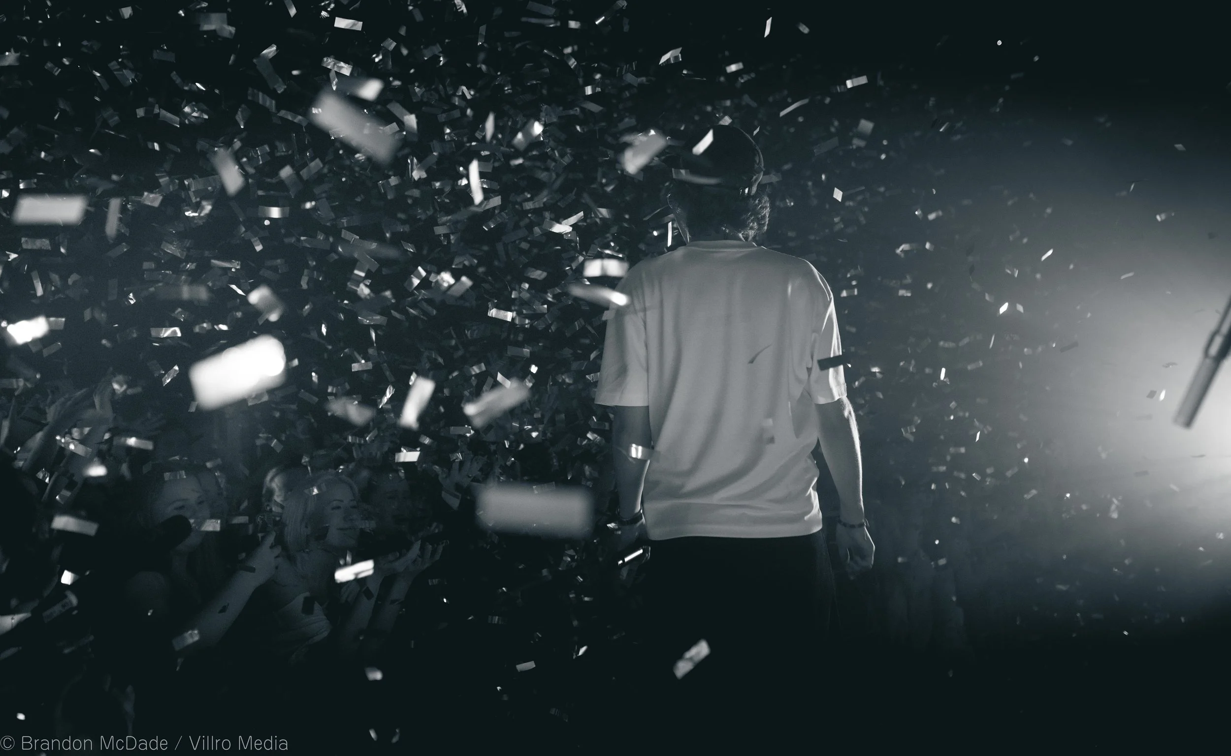 A person standing on a stage surrounded by falling confetti, facing an audience with cheering on their phones, in black and white.
