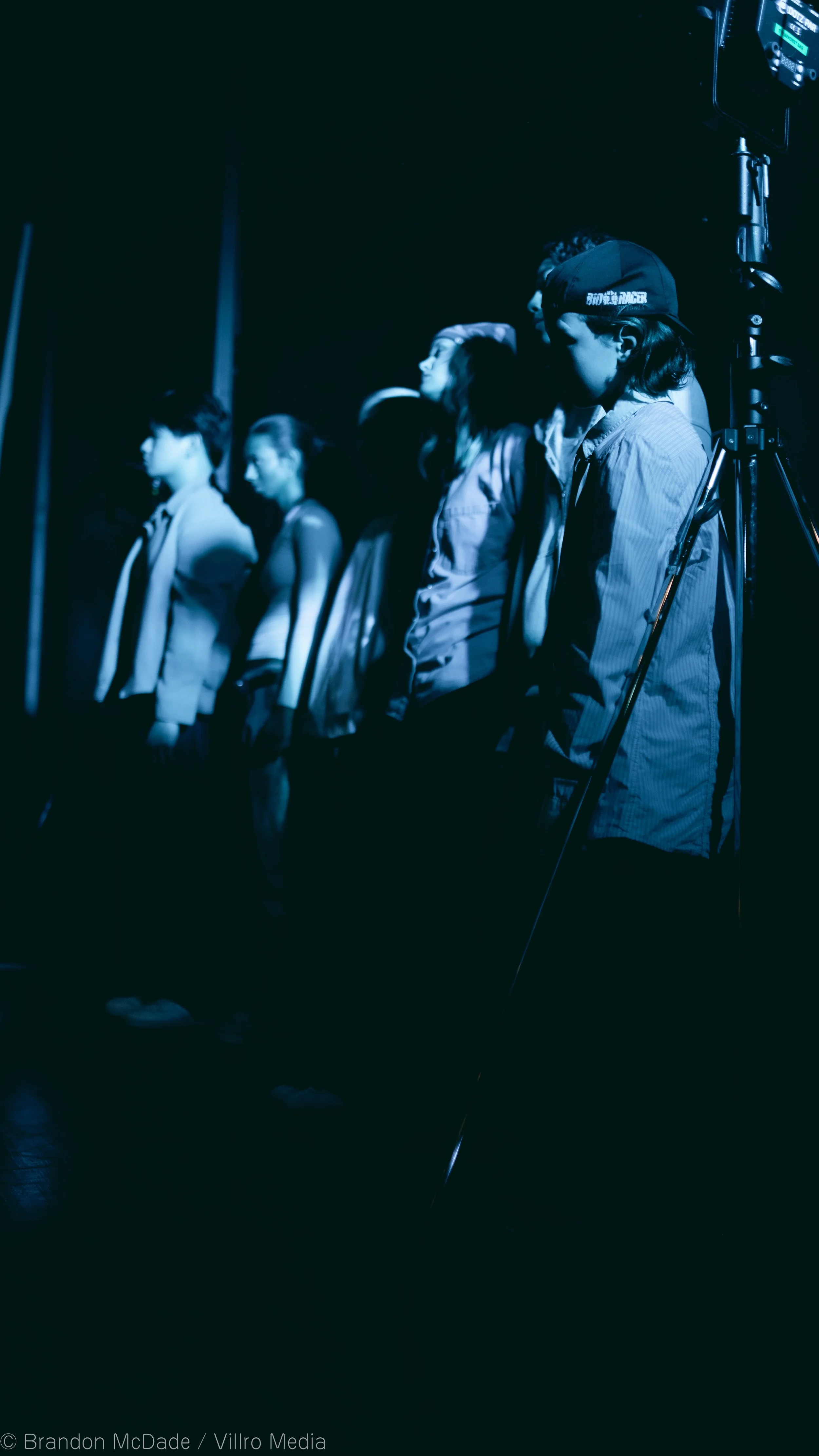 Group of people standing in a dark room, illuminated by blue stage lights, possibly at a concert or event.