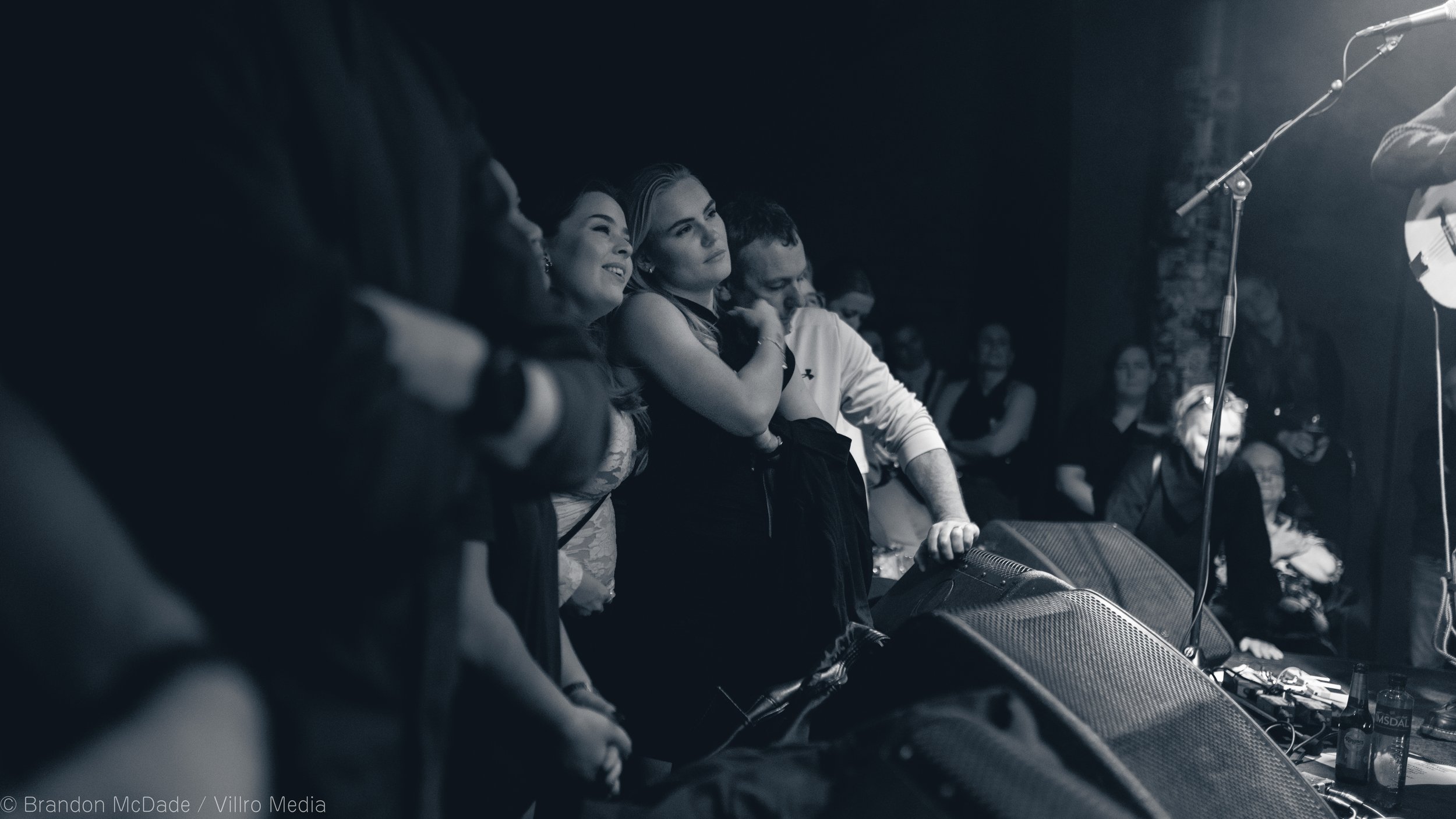 A group of people standing in front of a stage, with some of them smiling and watching a performance. The scene appears to be in a dark venue, possibly a concert or comedy show.