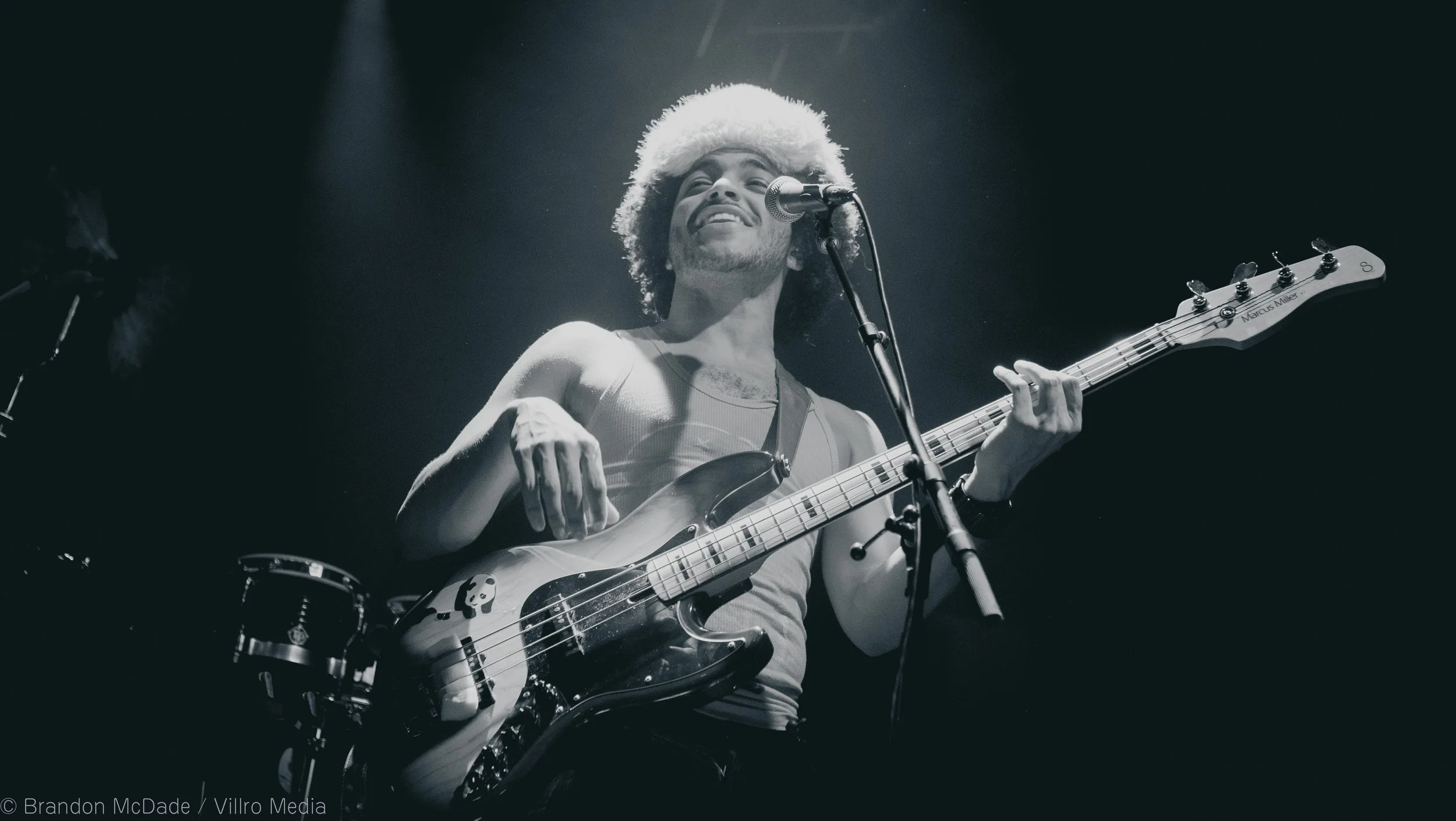 A man with curly hair wearing a tank top, playing an electric bass guitar and singing into a microphone on stage, illuminated by a spotlight.